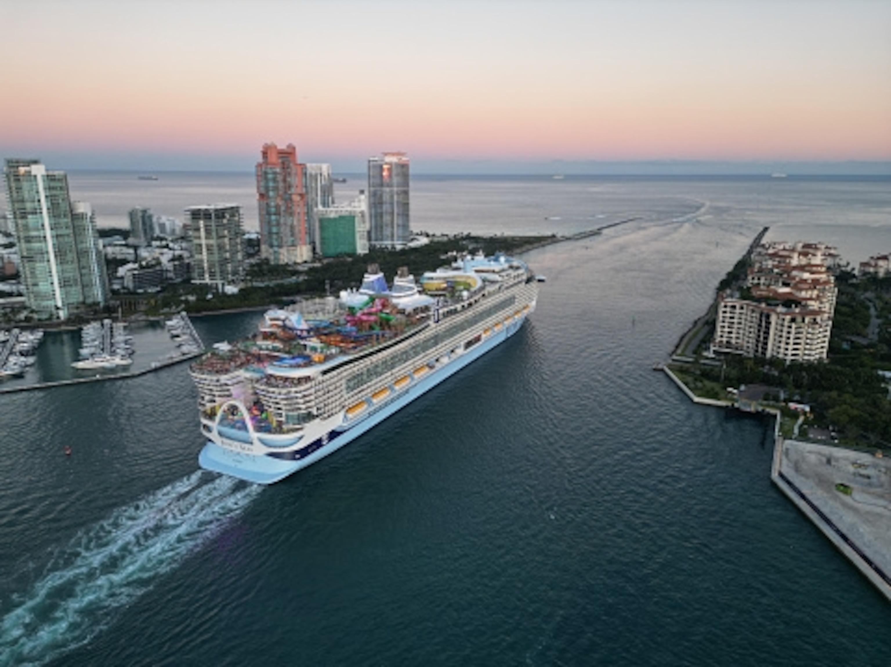 An enormous cuise ship sailing away from a coastal city at sunset. Skyscrapers and waterfront buildings line the shore.