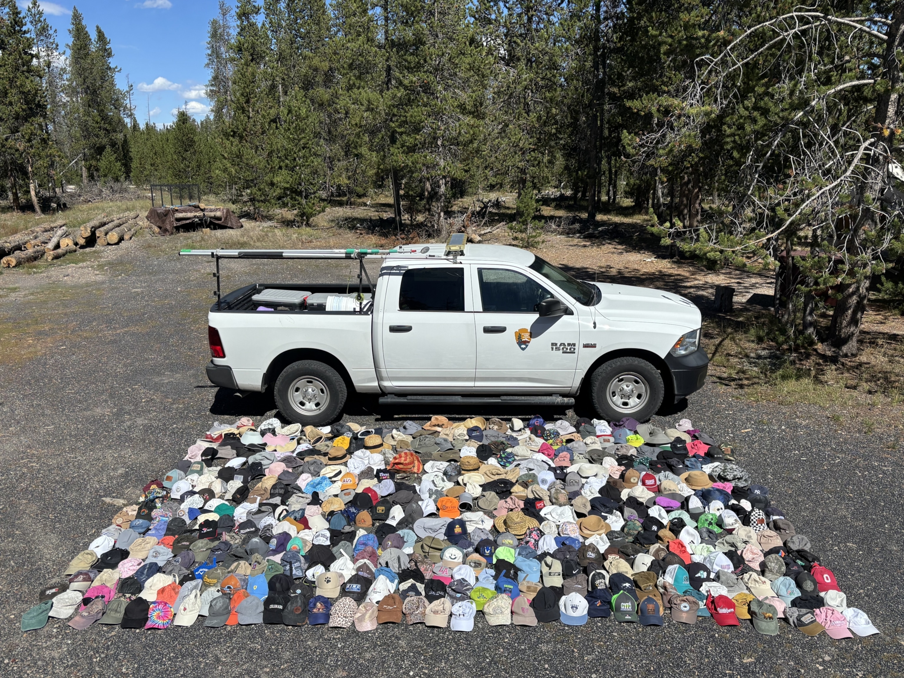 A white National Park Service pickup truck is parked in a forest clearing, with hundreds of hats and caps arranged on the ground in front of the vehicle in a large grid pattern.