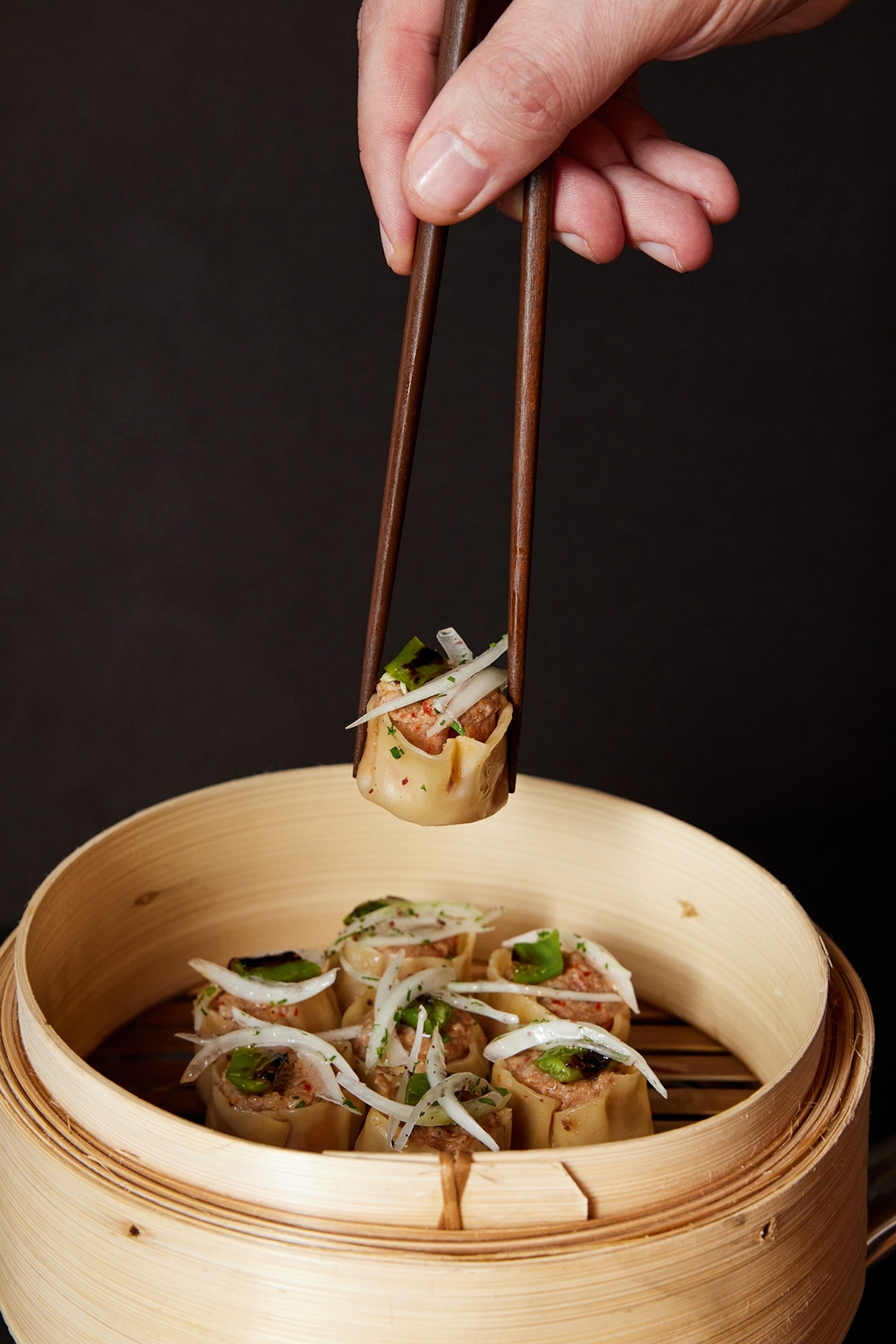 A food closeup of a wooden dim sum steamer holding a seven open-ended dumpling cups filled with meat as two chopsticks hover above with an eighth one.