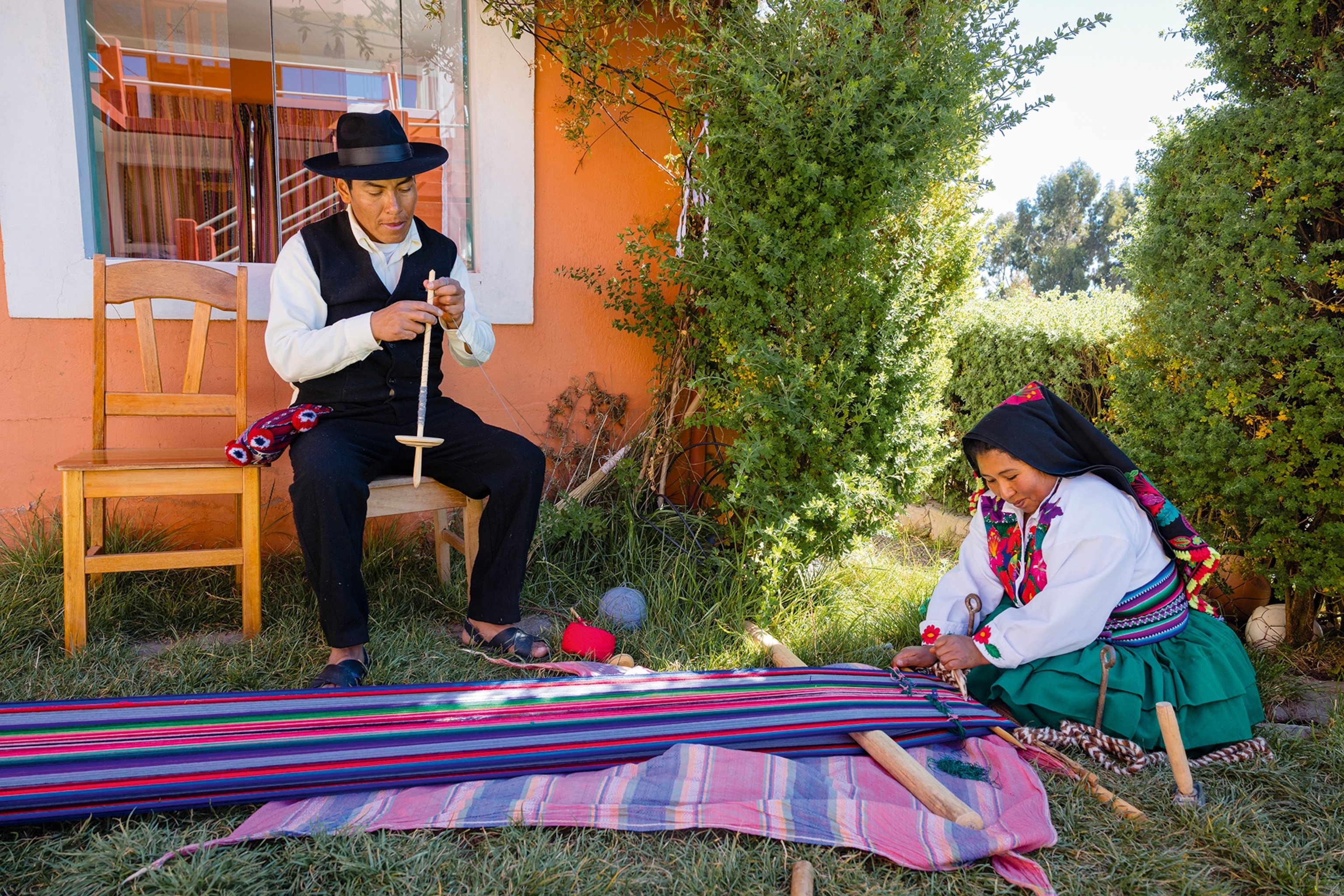 A native Peruvian couple in the garden in front of their house, the woman weaving a long piece of fabric on the grass, while her husband adjusts a wooden tool.