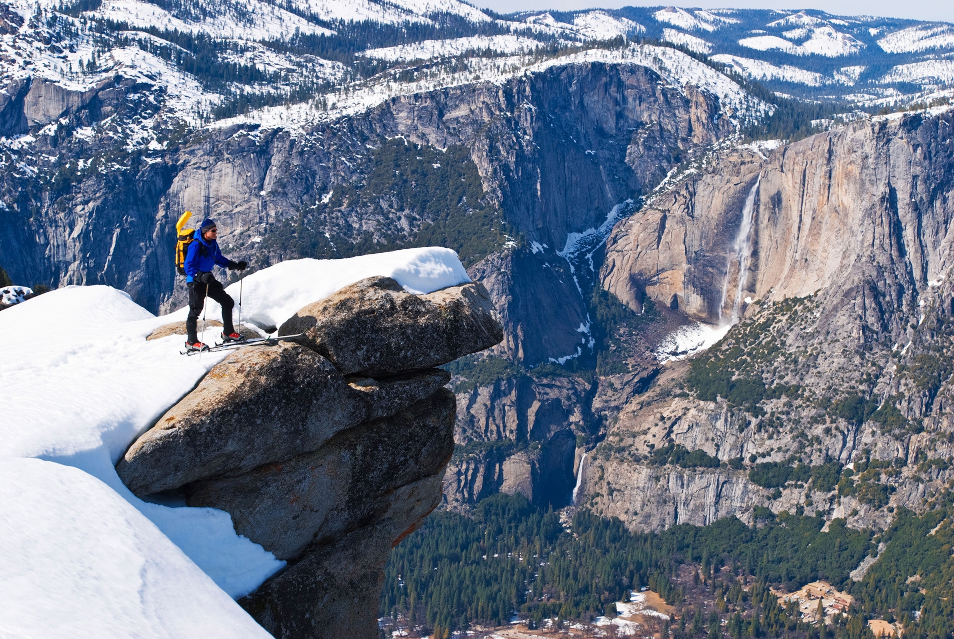 A wide shot of a massive, deep valley dusted in snow and with a waterfall. A man in a blue coat and on skis stands on a rocky outcropping looking out over the valley.