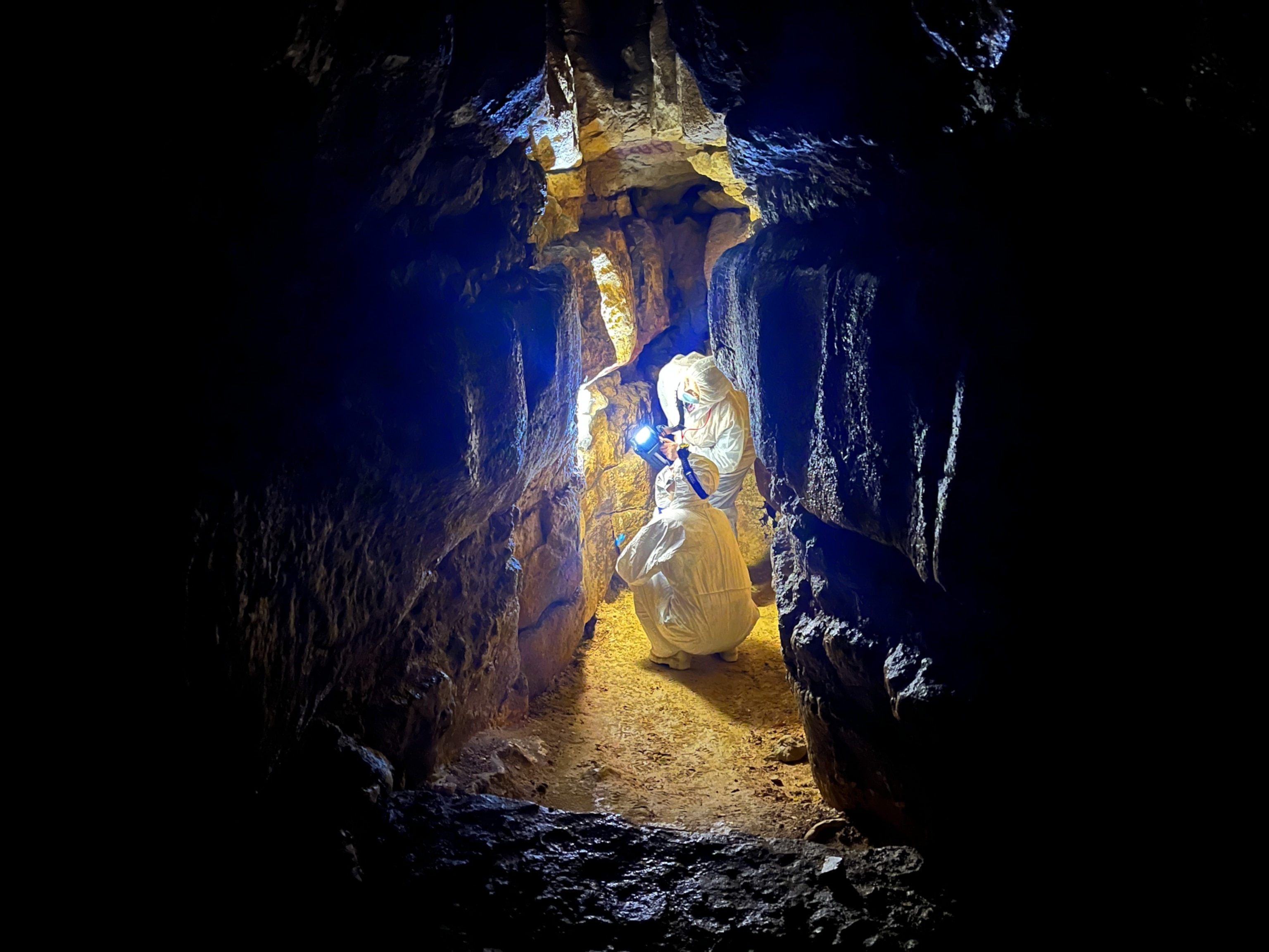 Two people in protective suits explore a narrow, dimly lit cave passage, holding a bright light