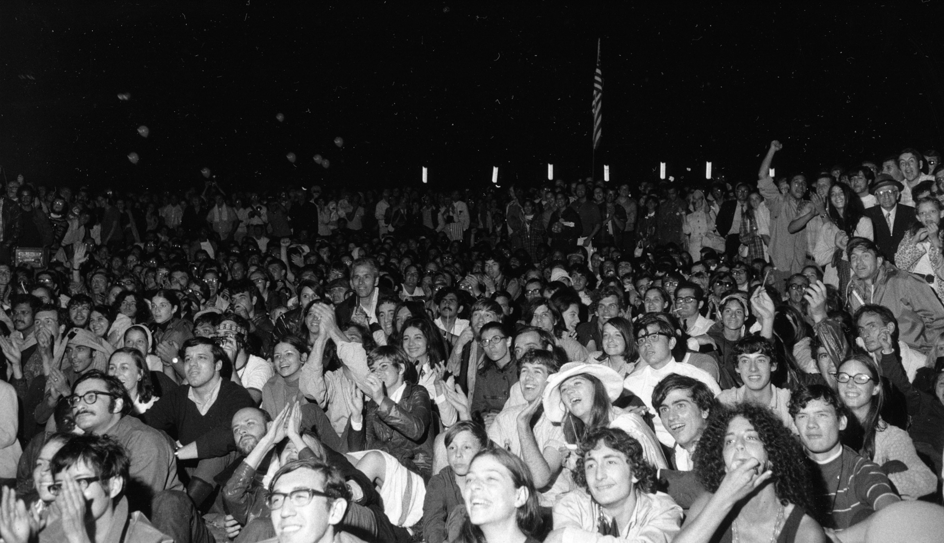 A large, diverse crowd sits closely together in a nighttime setting, many smiling and clapping