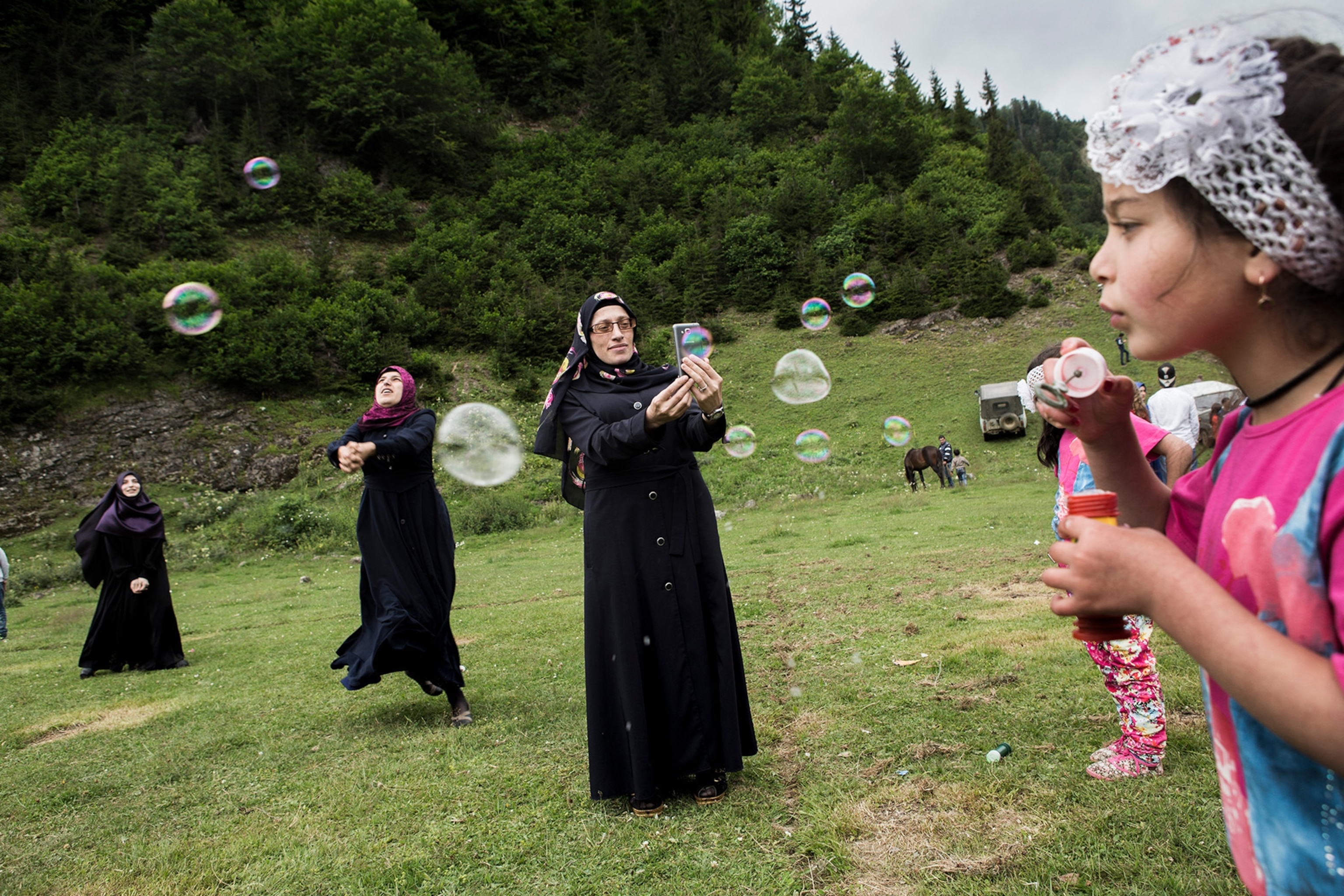 A woman wearing a hijab holds her phone up to a young girl blowing bubbles in a grassy area, while two other women with hijabs frolick behind her.
