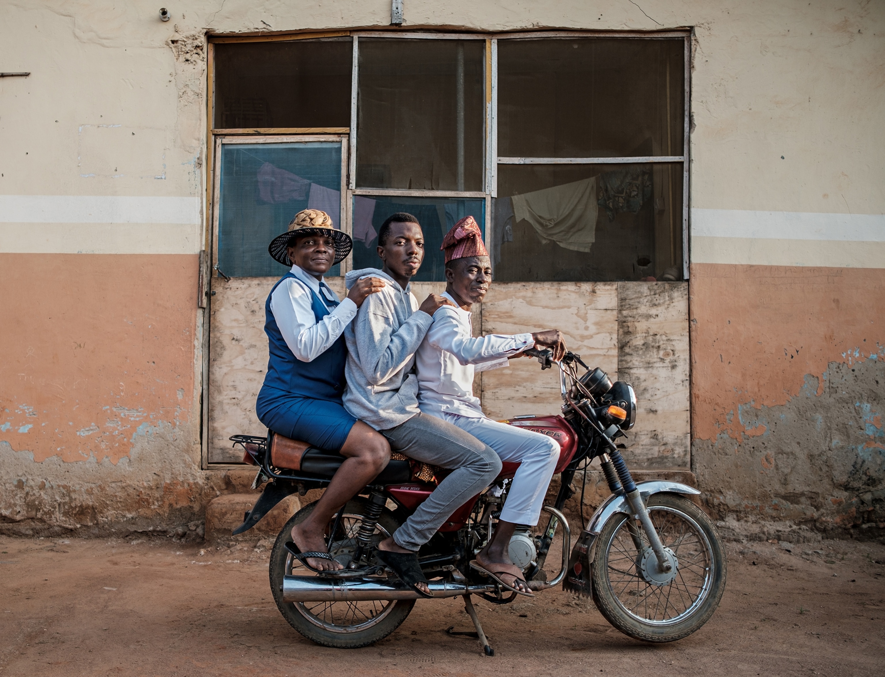 Two men and woman in blue dress and hat on the motorcycle looking into camera.