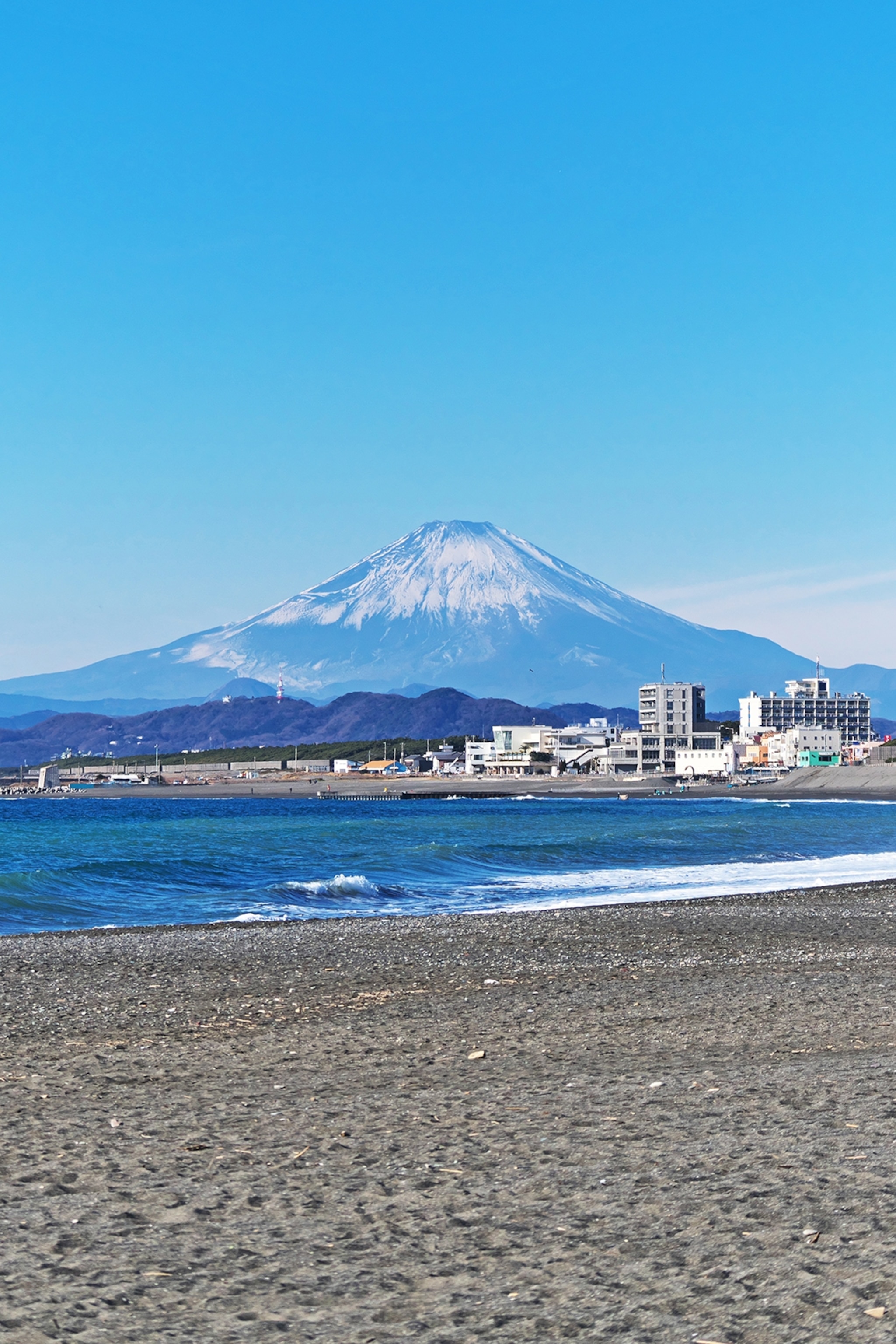 A gravel beach with big, wide waves crashing on the shore outside a small town, Mount Fuji is seen in the distance.