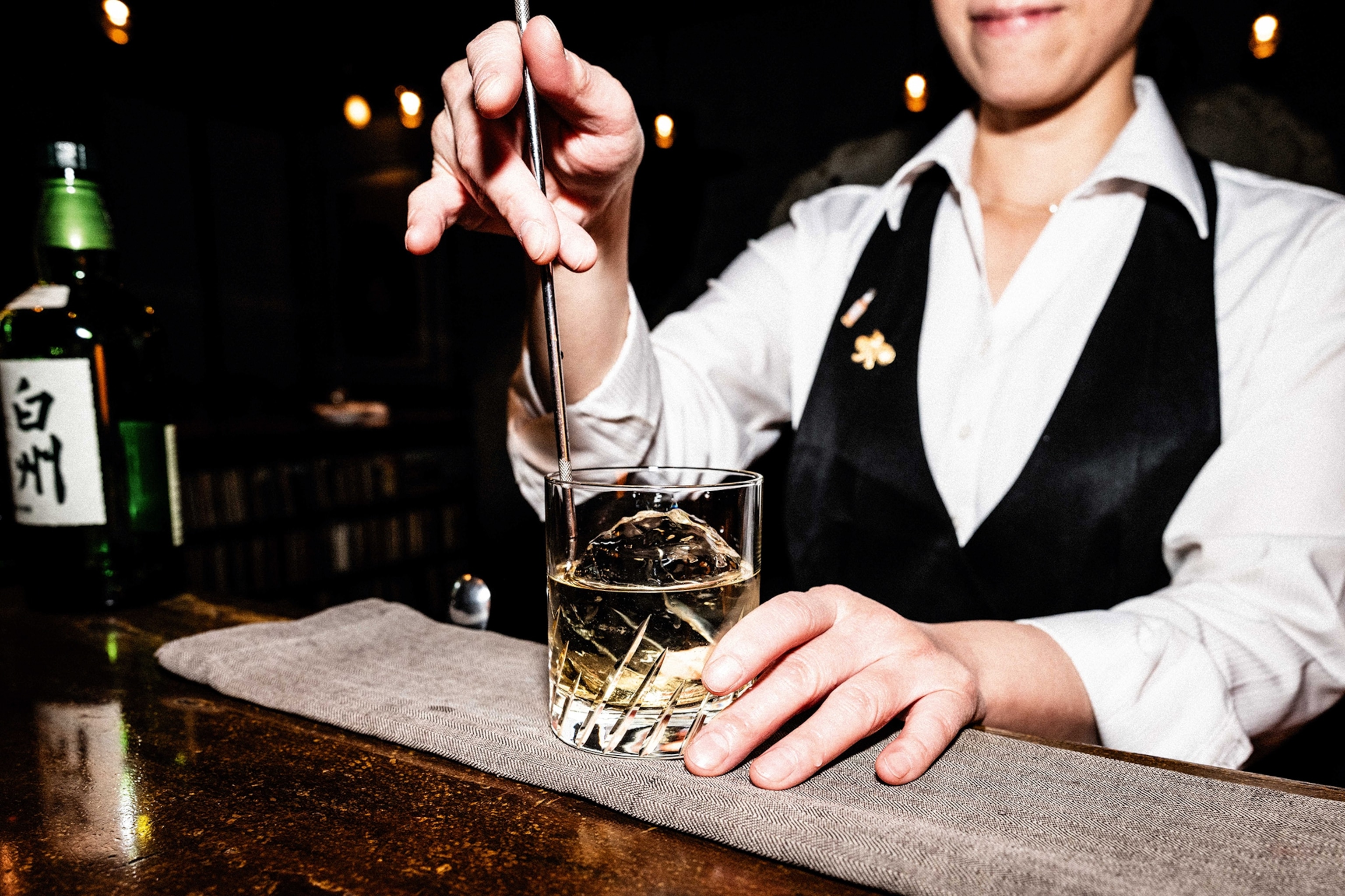 A female bartender stirring a tumbler of Japanese whiskey with ball-shaped ice cube on the top of a bar.