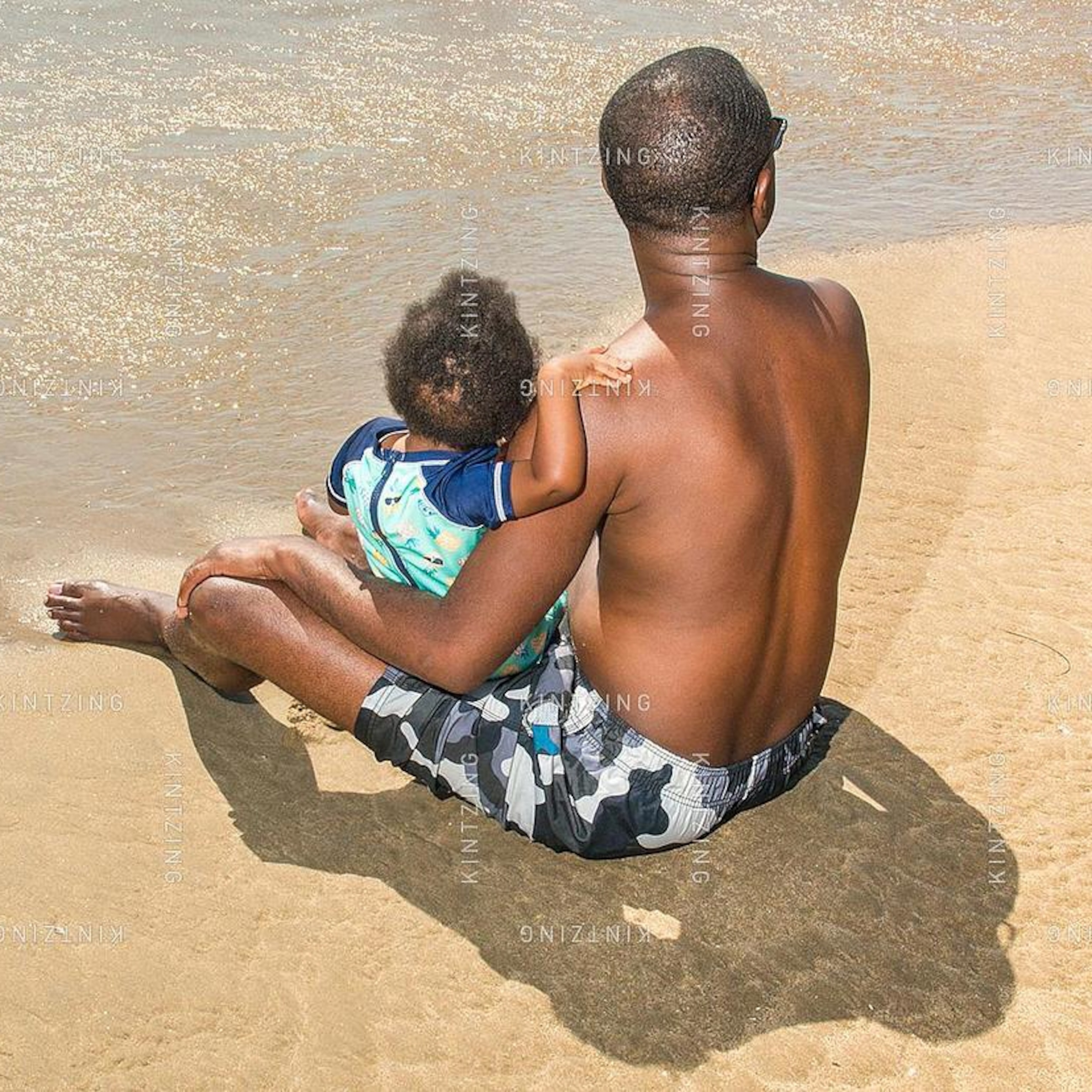 A father and child are seen from behind, sitting together on a sandy beach as they enjoy the view of the water.