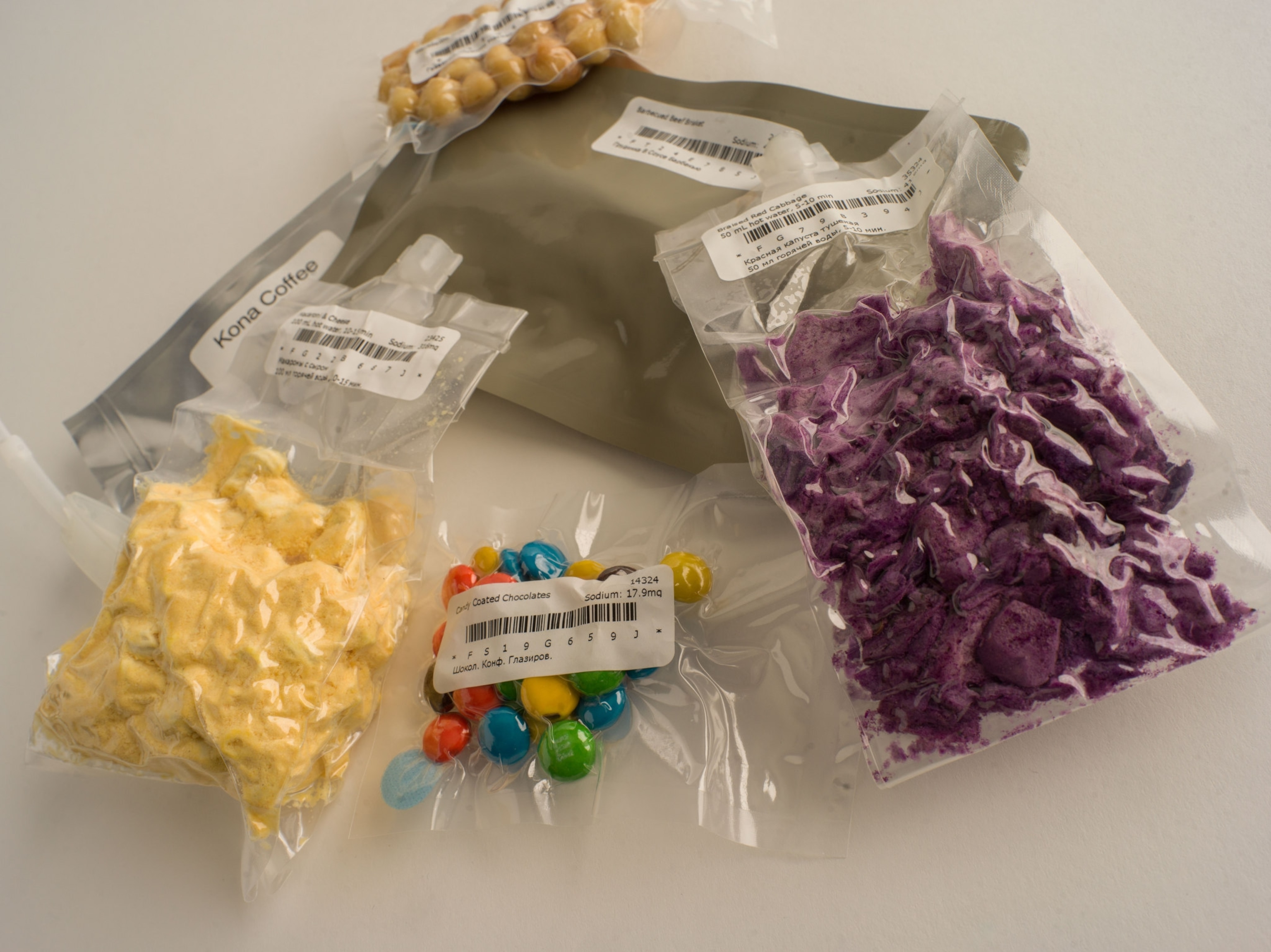Packaged NASA food on table- Kona coffee, macadamia nuts, beef brisket, candy coated chocolates, macaroni and cheese, braised red cabbage, kona black coffee.