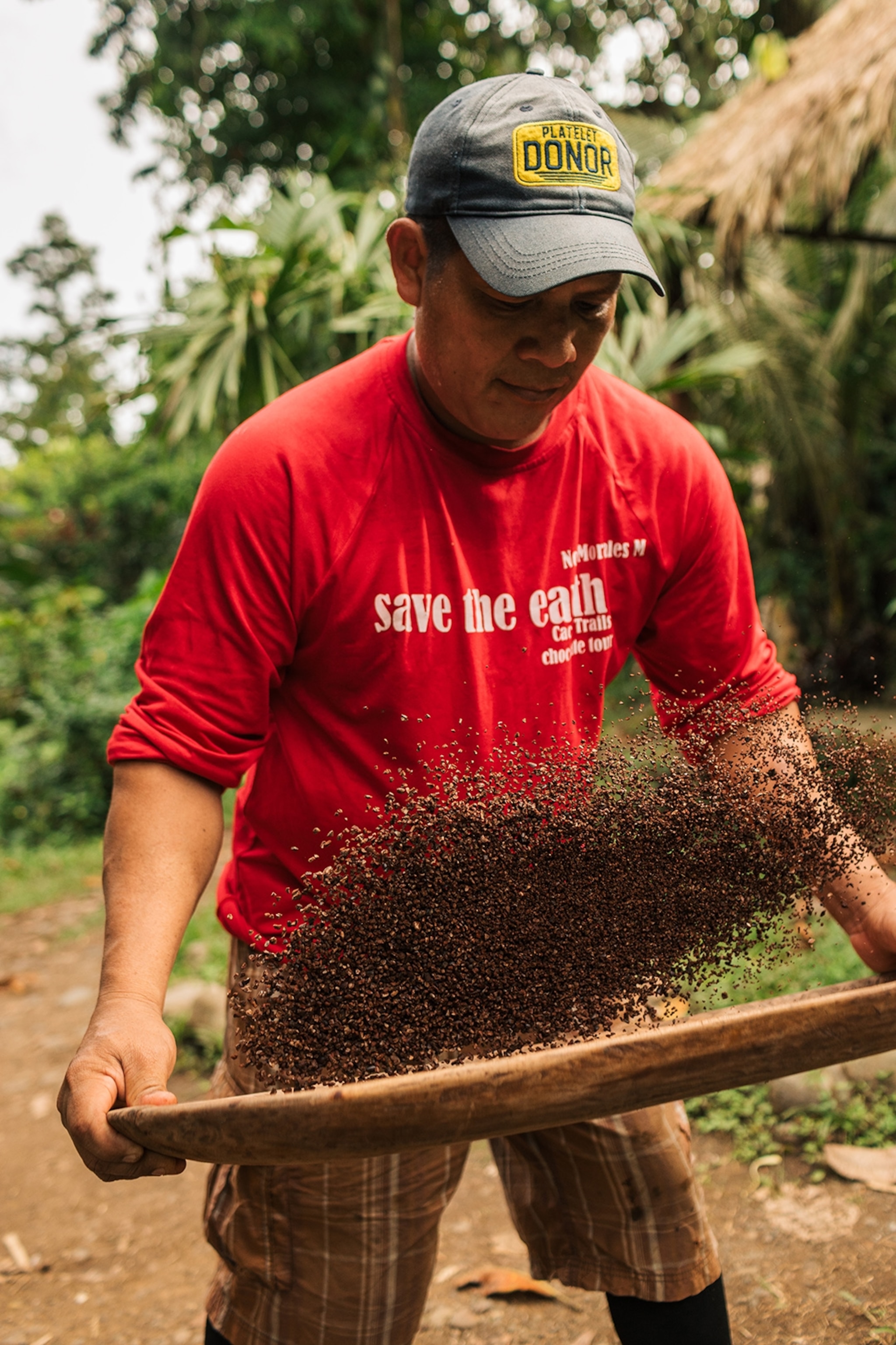 A male farmer in a simple t-shirt, shaking cocoa nibs in a straw sieve.