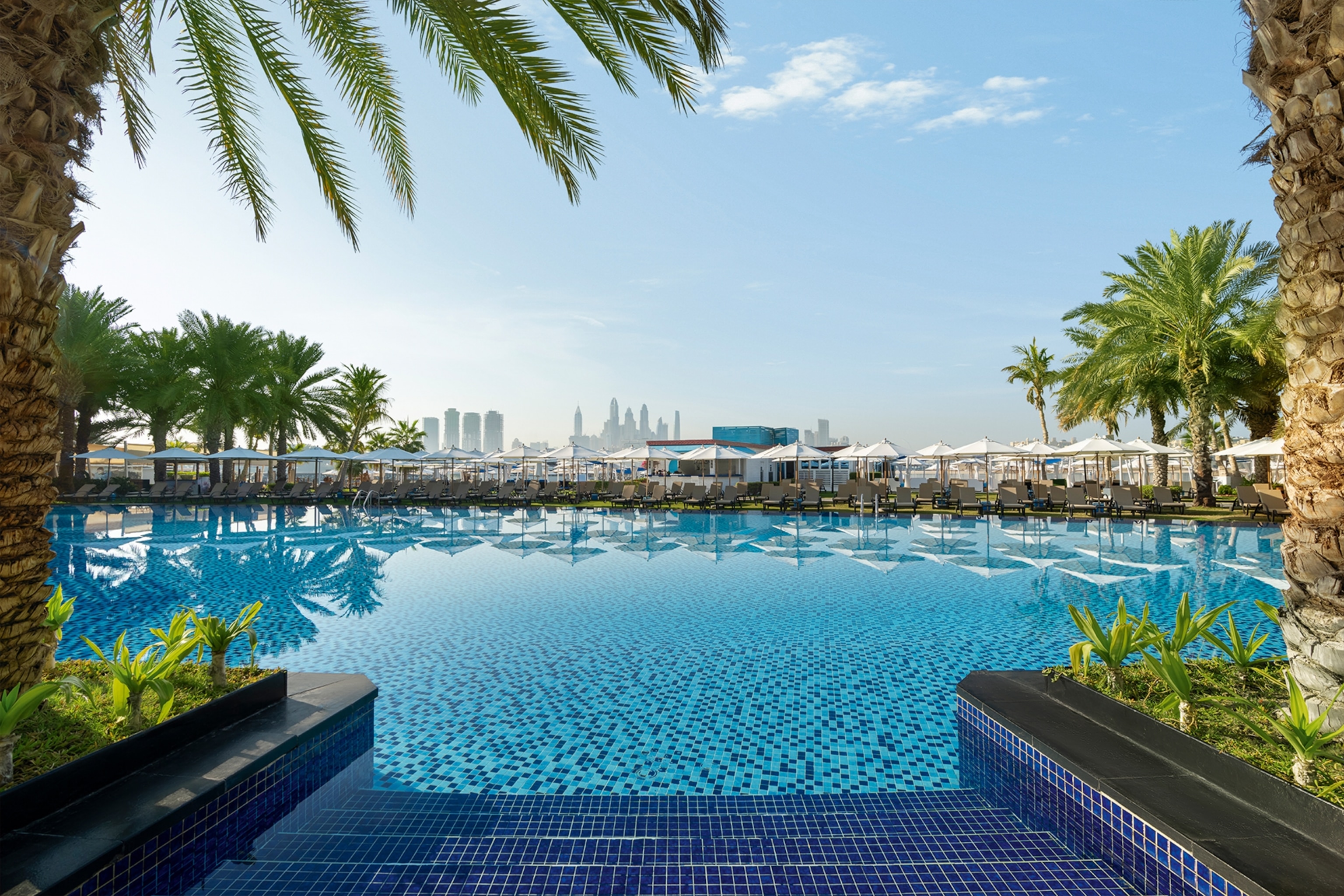 Stairs leading into a wide pool with sunchairs and parasols in front of a city skyline in the distance.