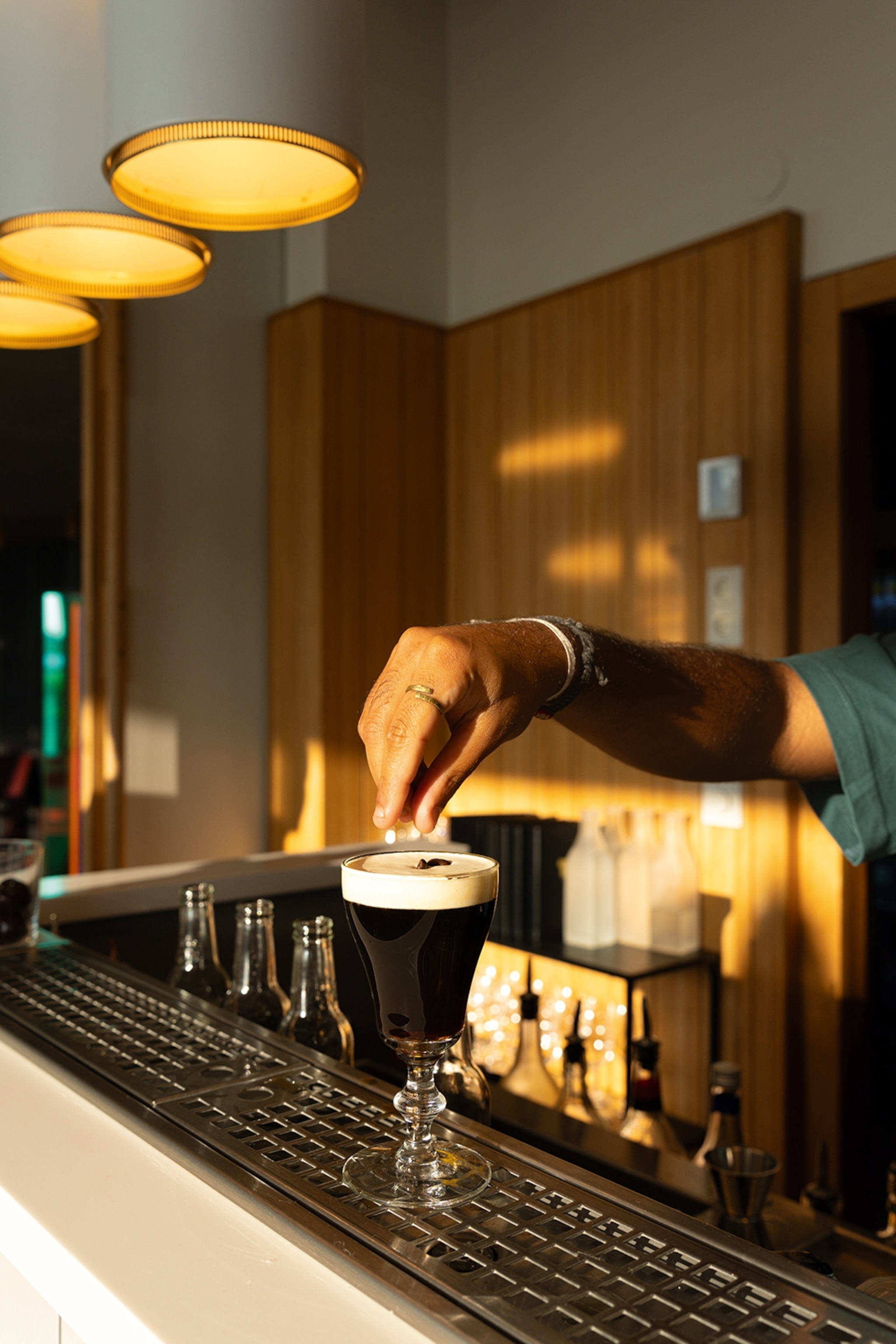 A close-up of a bar counter with an espresso martini and a hand dropping coffee beans onto the foamy top.