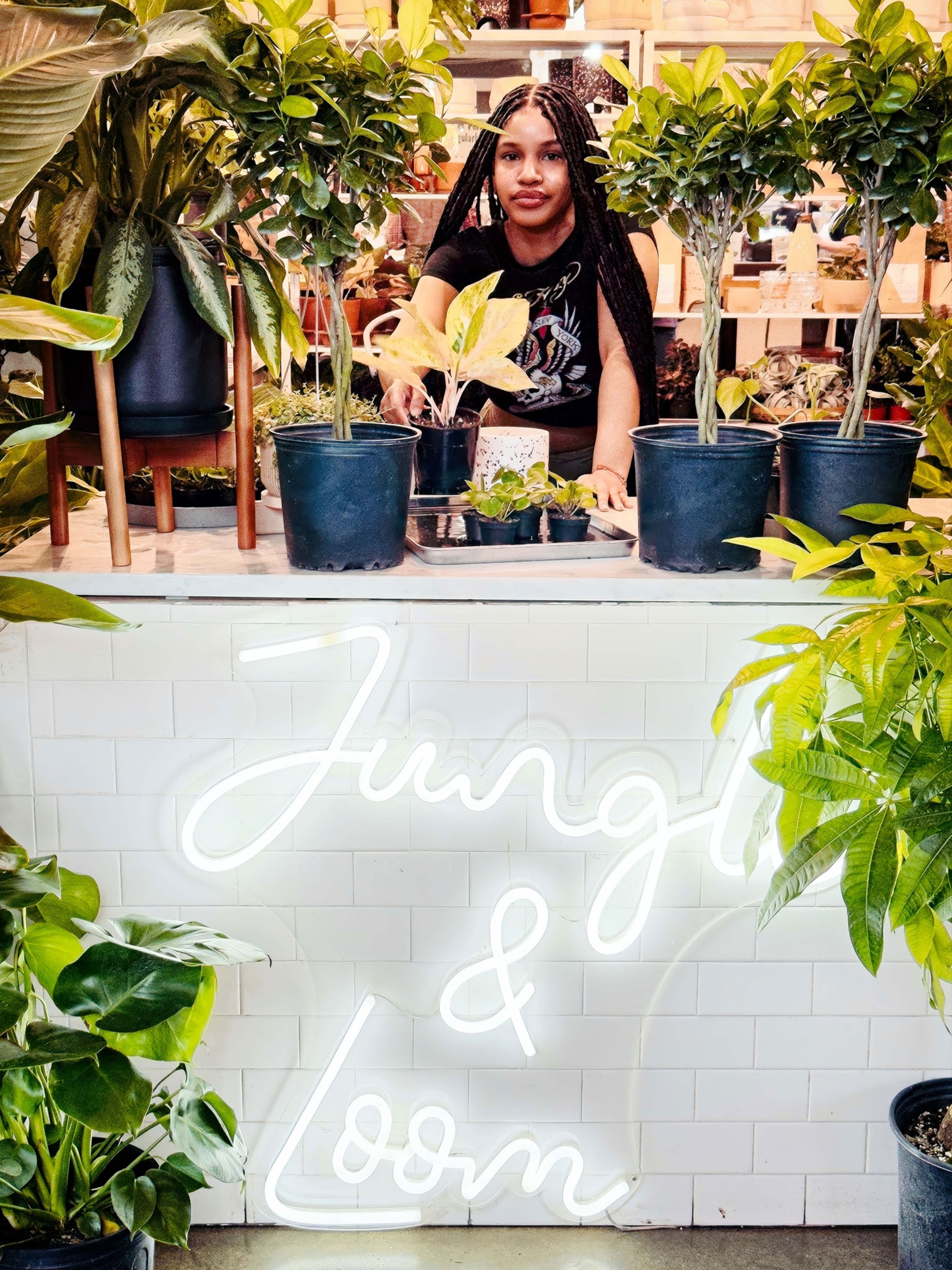 A woman with long box braids and a warm skitnone stands behind a white countertop, with a sign that has glowing letters.