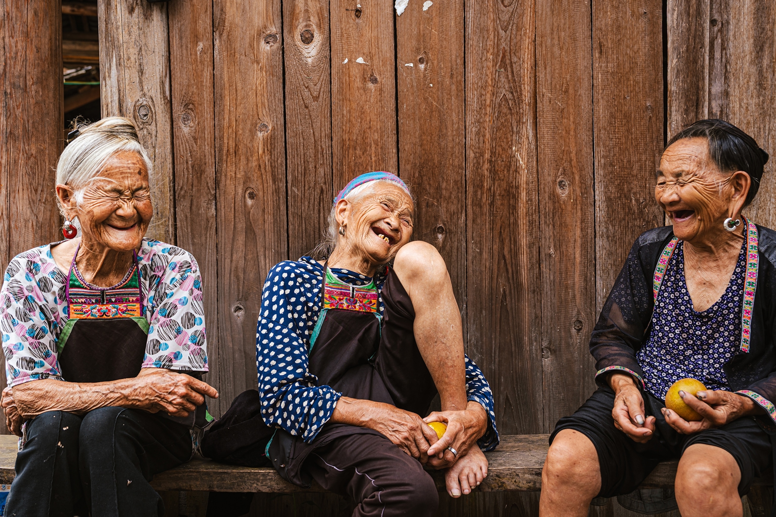 A group of three elderly Chinese women sitting on a wooden bench attached to a house and laughing widely.