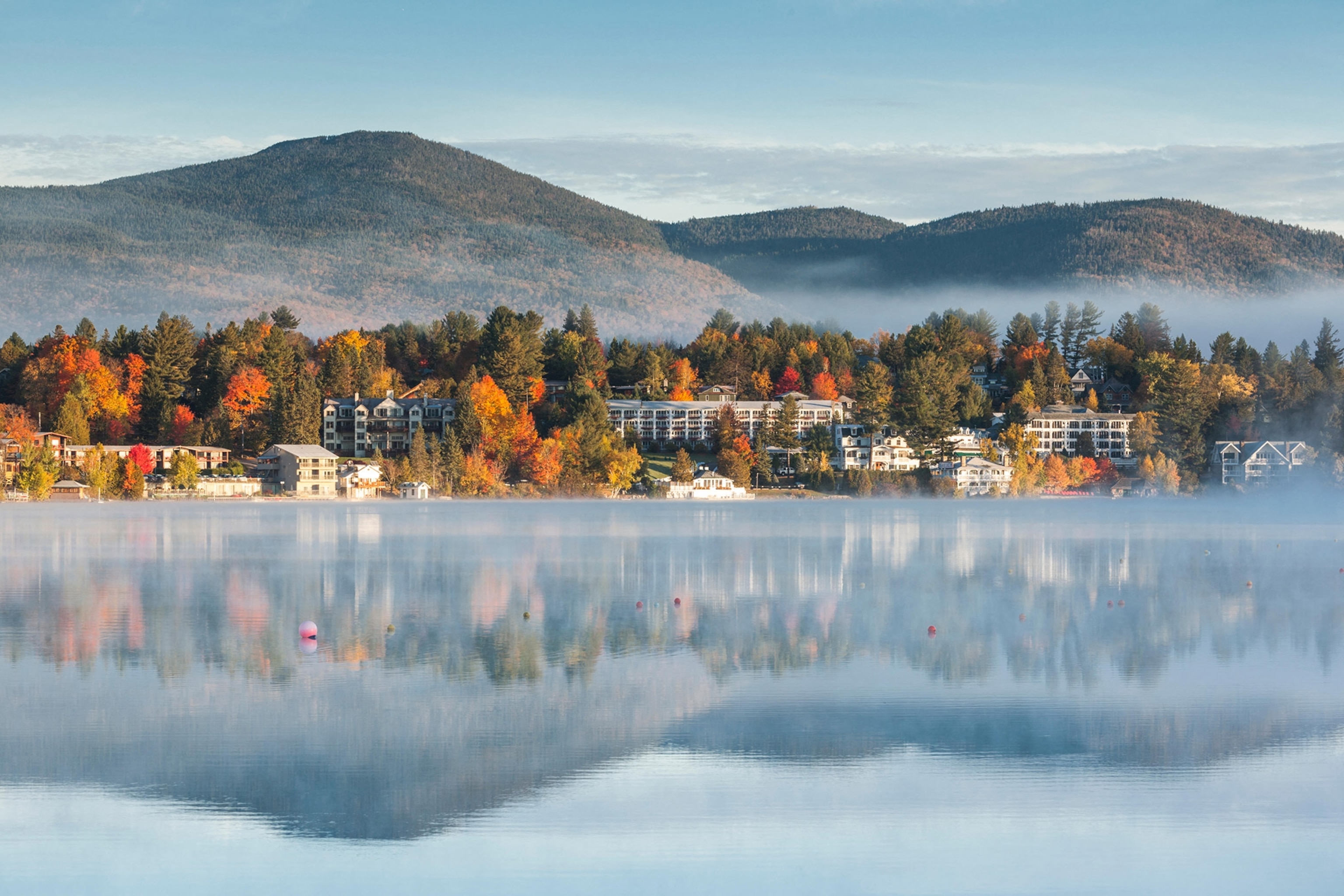 A village tucked among colorful trees and at the base of a mountain is reflected in a glassy lake