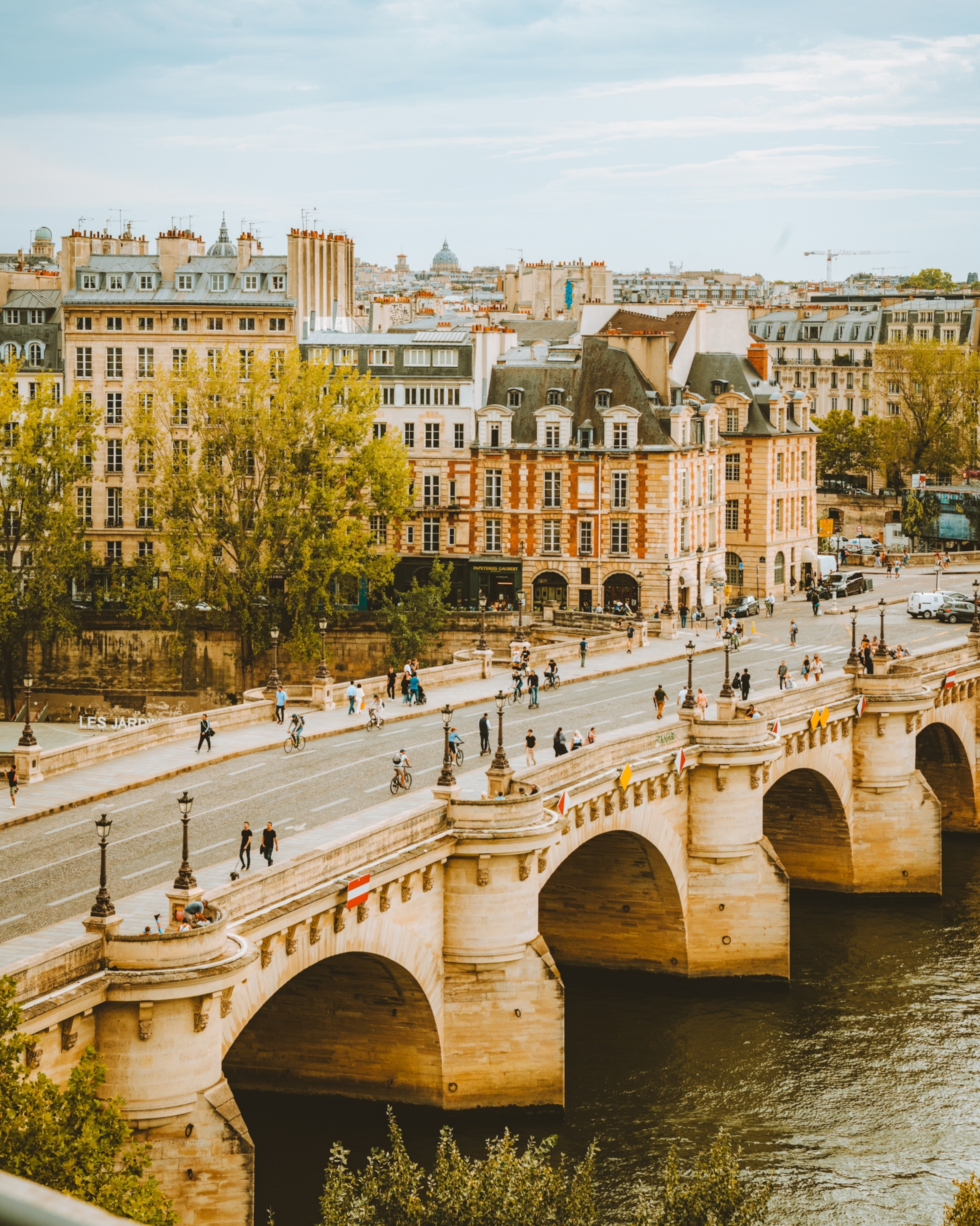 Pont Neuf, a stone bridge, spans the Seine River in Paris, with historic buildings, tree-lined streets, and people walking and cycling across the bridge.