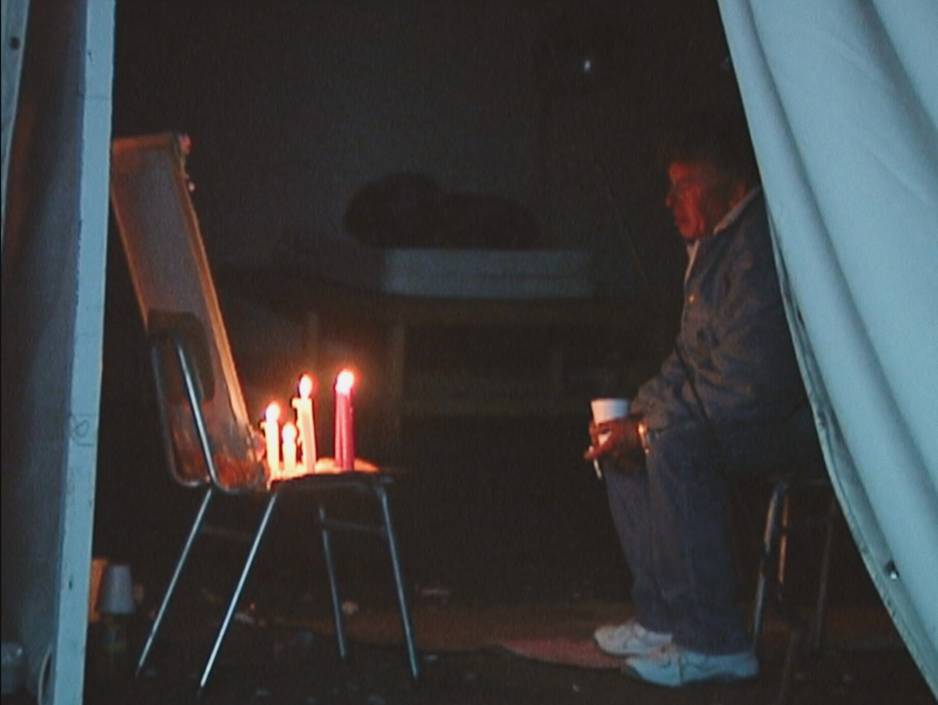 A man sits in a chair in a dark tent looking at a makeshift alter with candles burning on a chair.