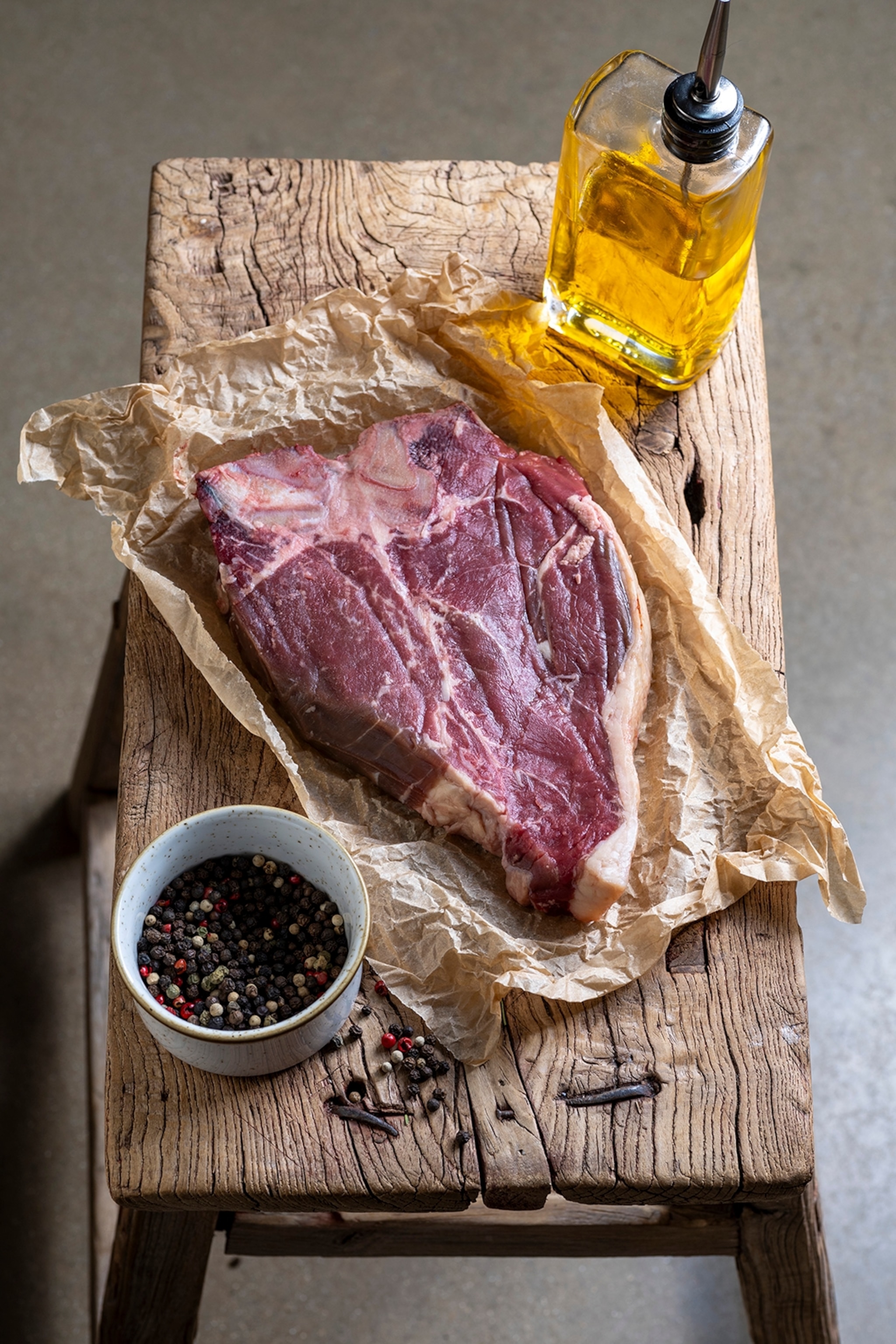 A rustic wooden chopping board with a piece of marble raw meat alongside an oil caraffe.