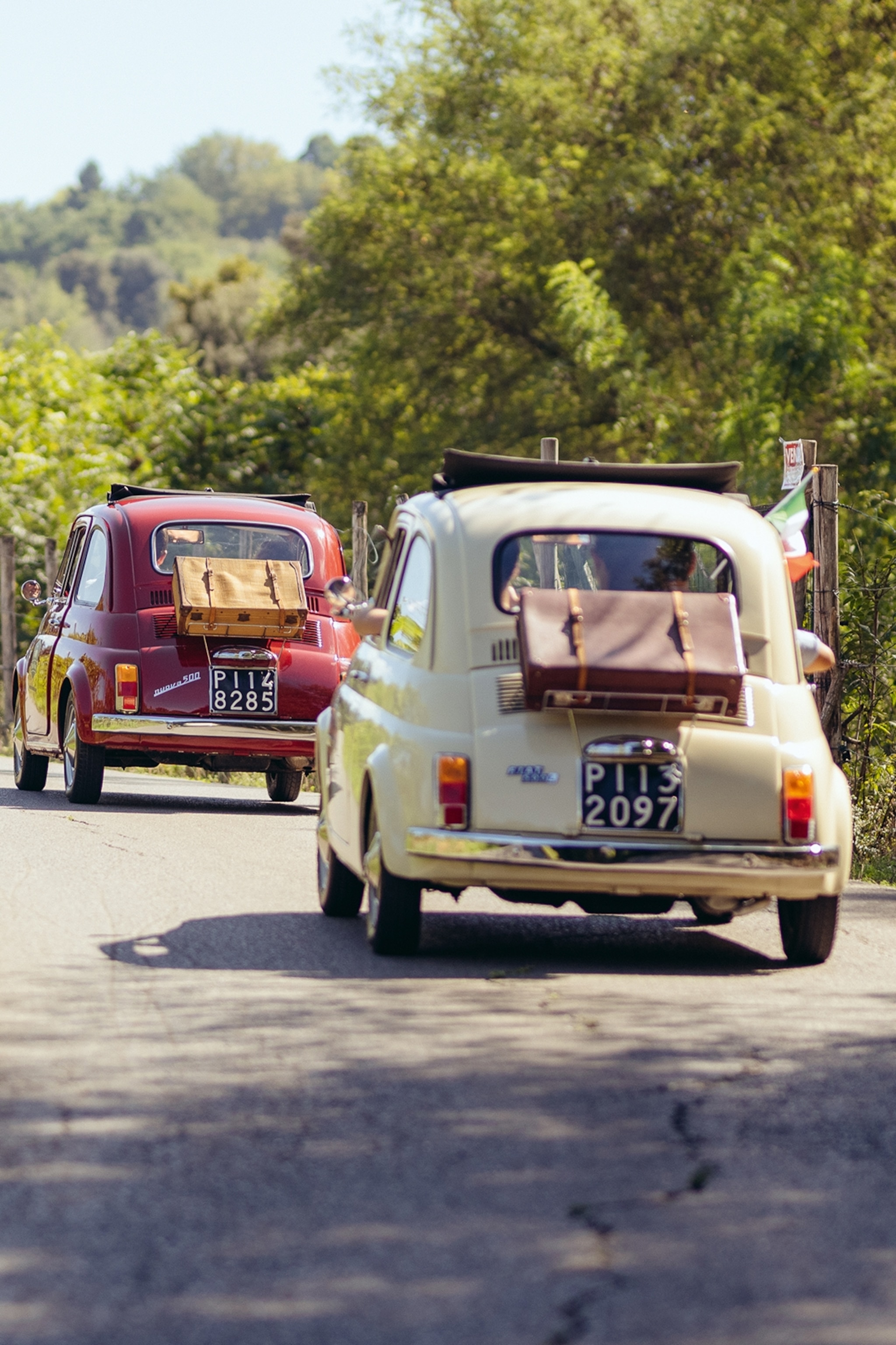 Two retro Fiat 500's making their way up a concrete road leading across a hill through lush forests.