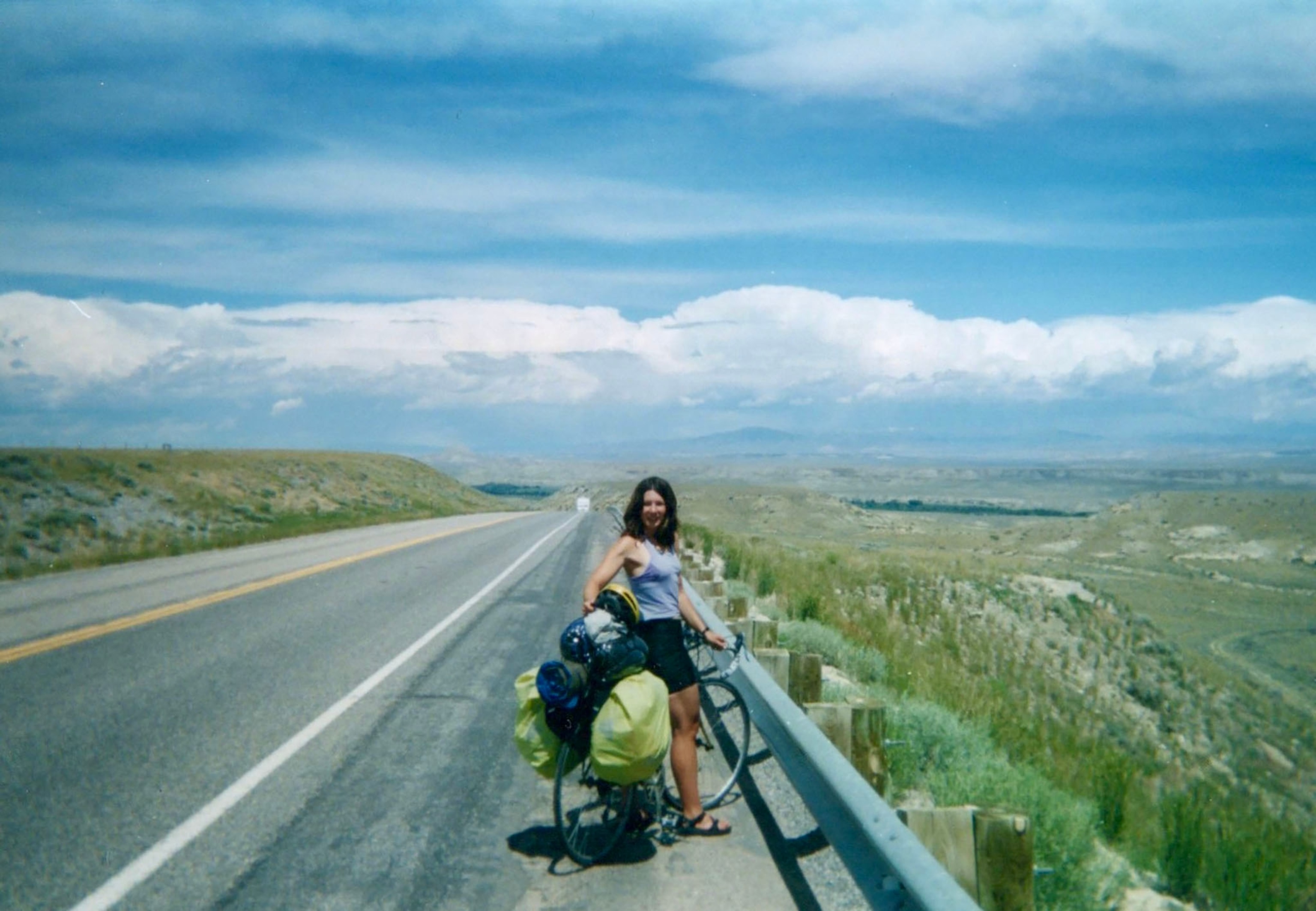 A woman stands with her bike, looking back at the camera, on the road under a big blue sky.