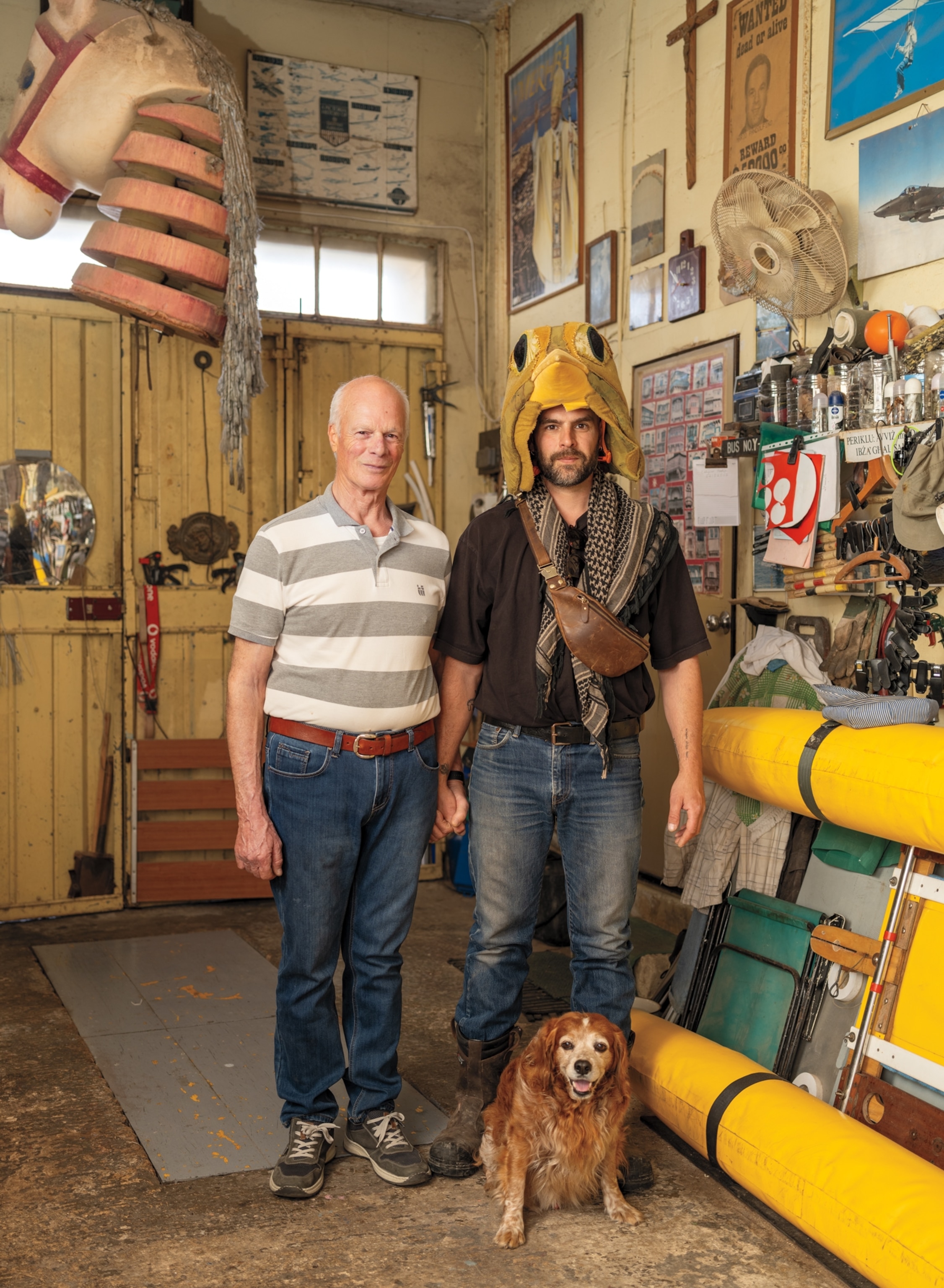 An older man in a gray-and-white striped shirt and jeans holds hands with a younger man in a yellow hat, black shirt, scarf, and jeans. A small dog sits in front of them.