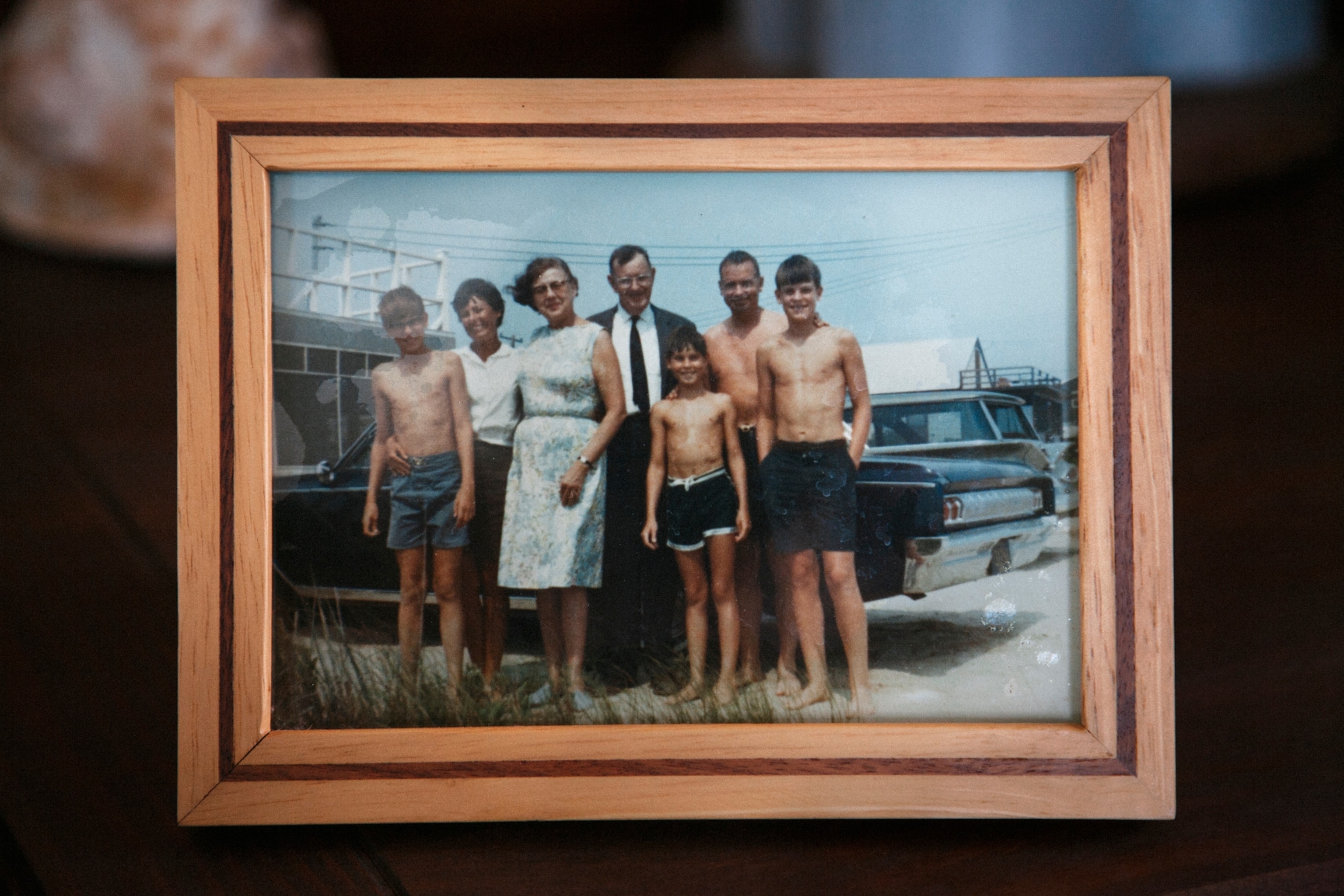 an old family photograph in a wooden frame, with seven people, some young boys in beach wear and some adults in dress clothes, standing in front of a car