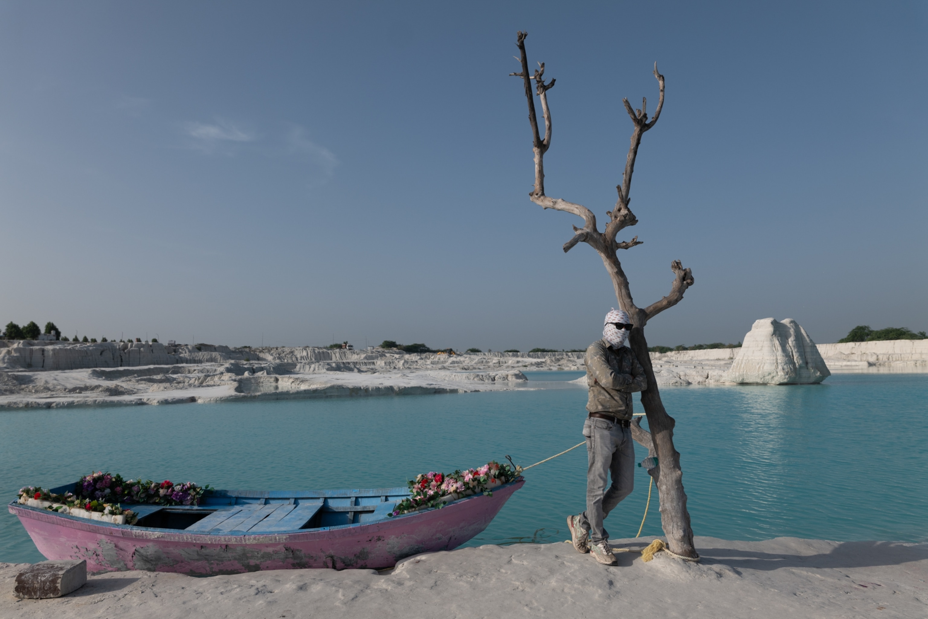 A man stands leaning on a tree, next too a pink boat on the water.