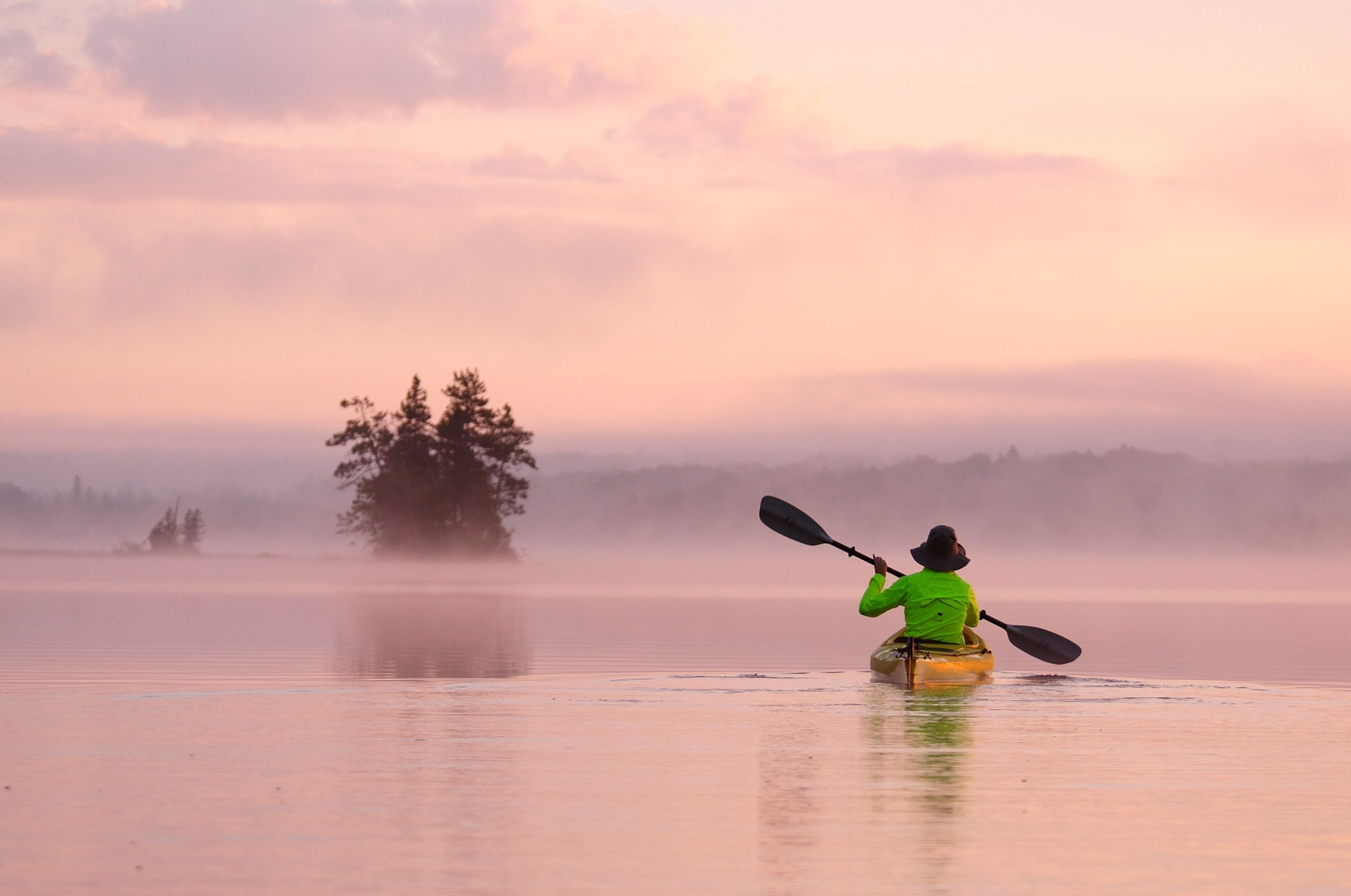 A woman in a yellow canoe with a green shirt kayaks into a pink sunset on a lake with trees in the distance