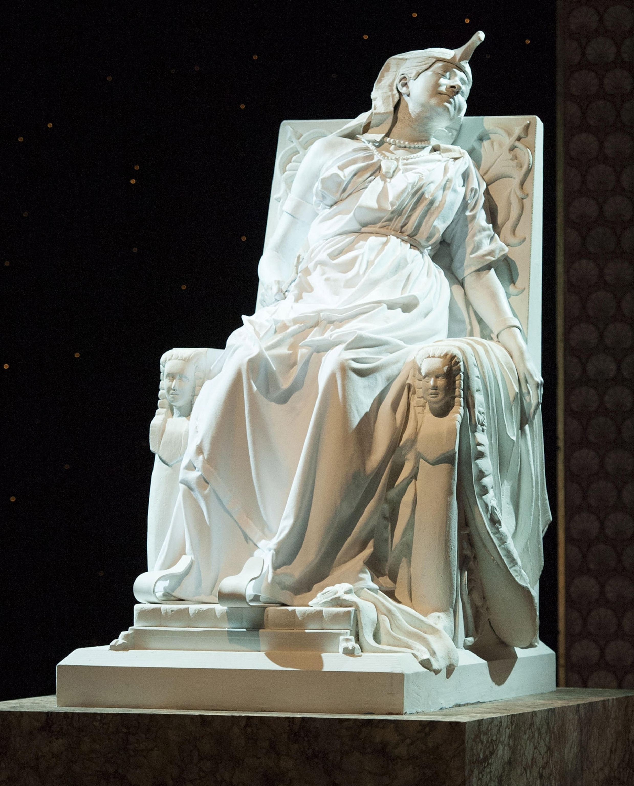 White marble sculpture of a woman in draped robes and a crown reclining on a throne
