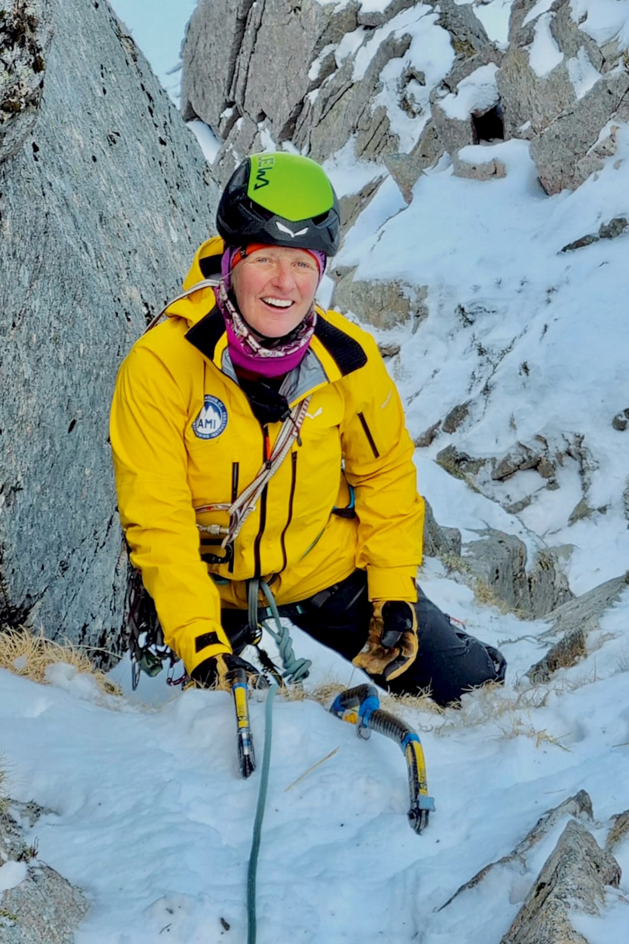 A smiling, mountaineering woman in between a snowy mountain ridge.