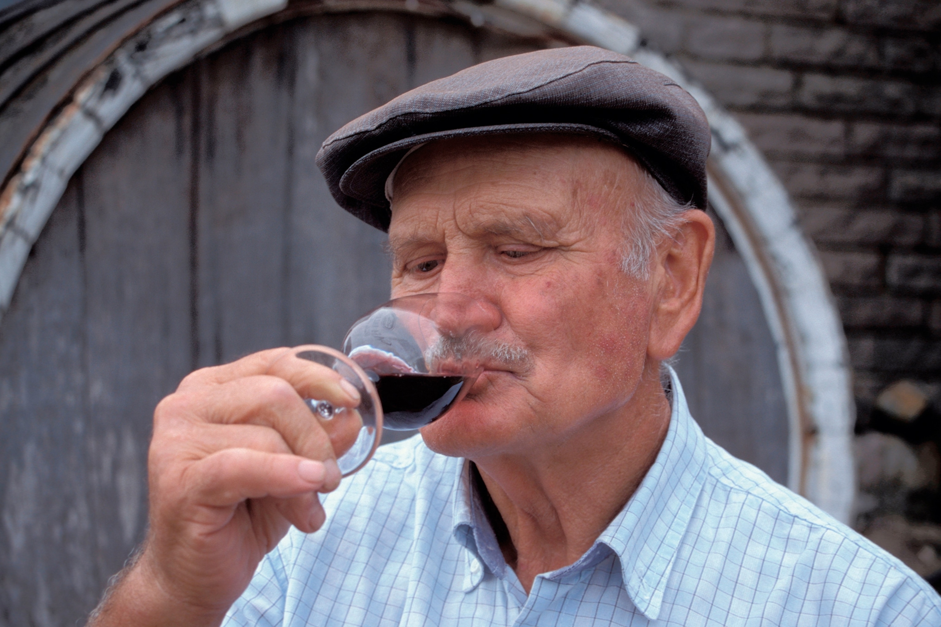 An elderly man with a flat cap and moustache sipping red wine from a glass.