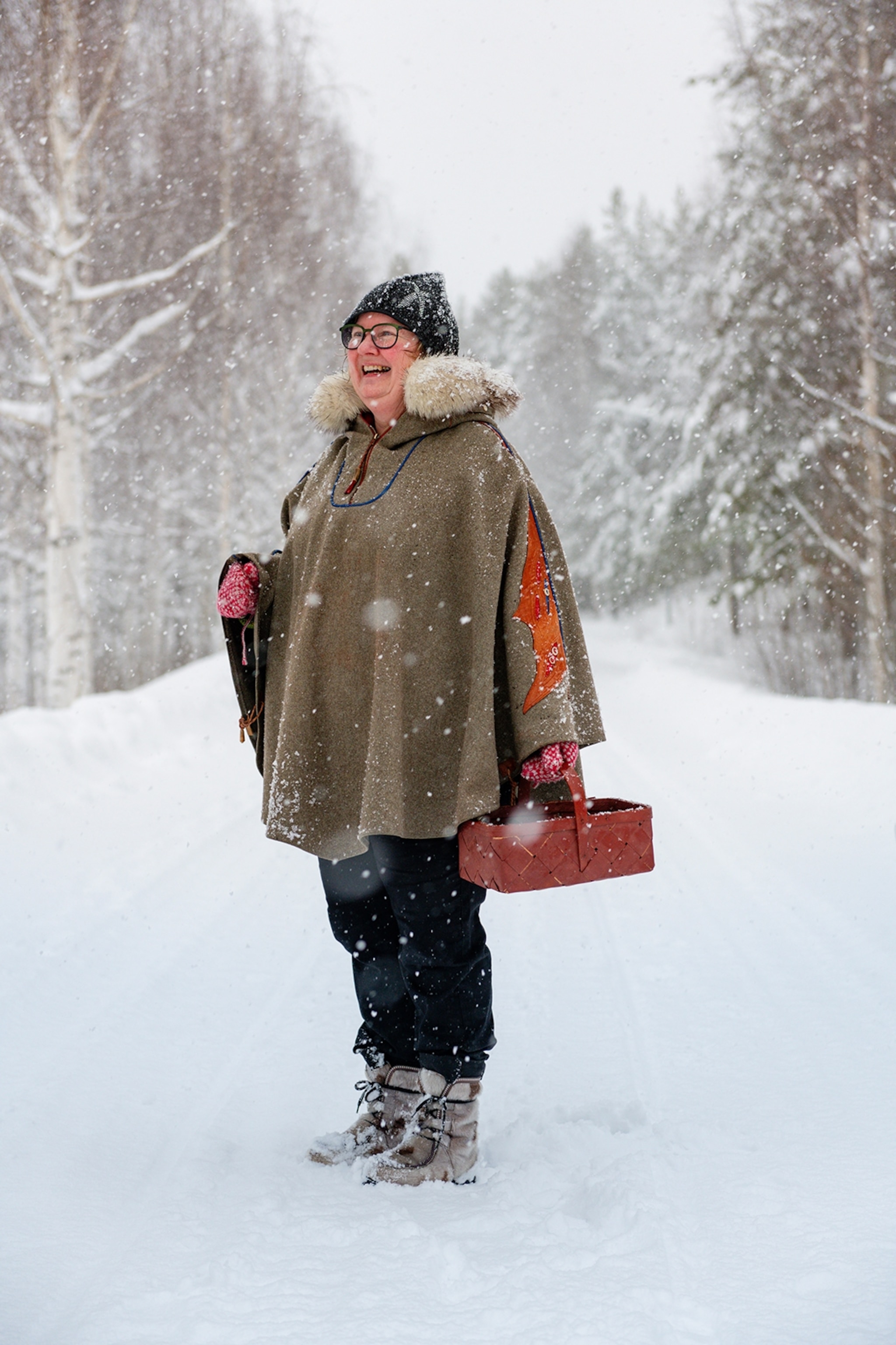 A smiling woman wearing a fur-necked winter cape in a snowy forest.