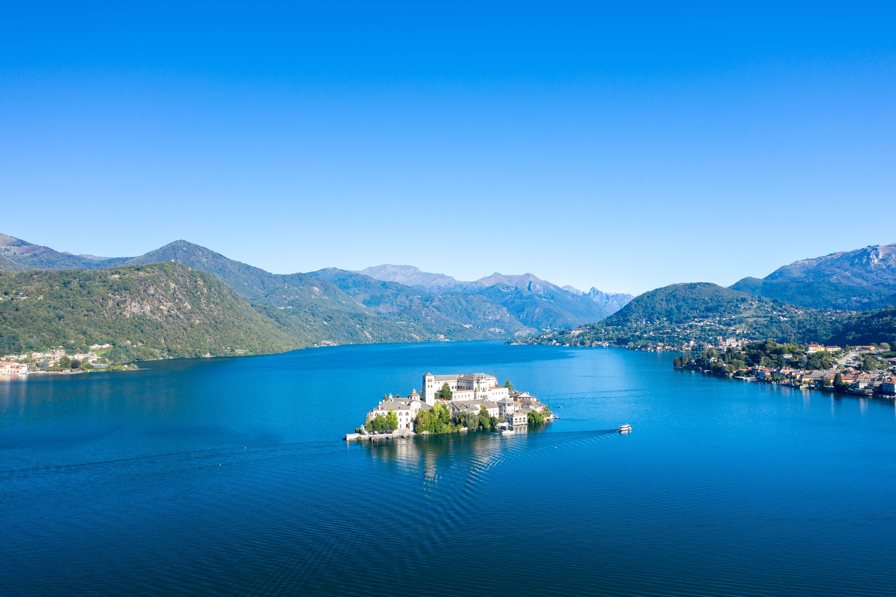 Aerial perspective of San Giulio Island in Lake Orta, northern Italy, encircled by deep blue water and framed by forested hills and distant alpine peaks. The scene features historic architecture and a tranquil natural setting beneath a vivid clear sky.