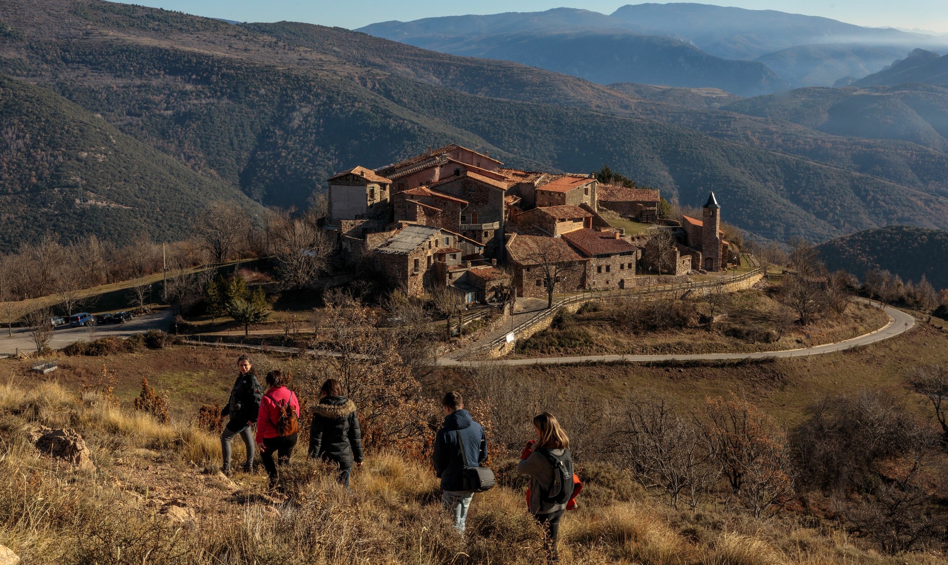 A small group of buildings gathered in a circle. Hikers head towards it, mountains are stretched out in the distance.