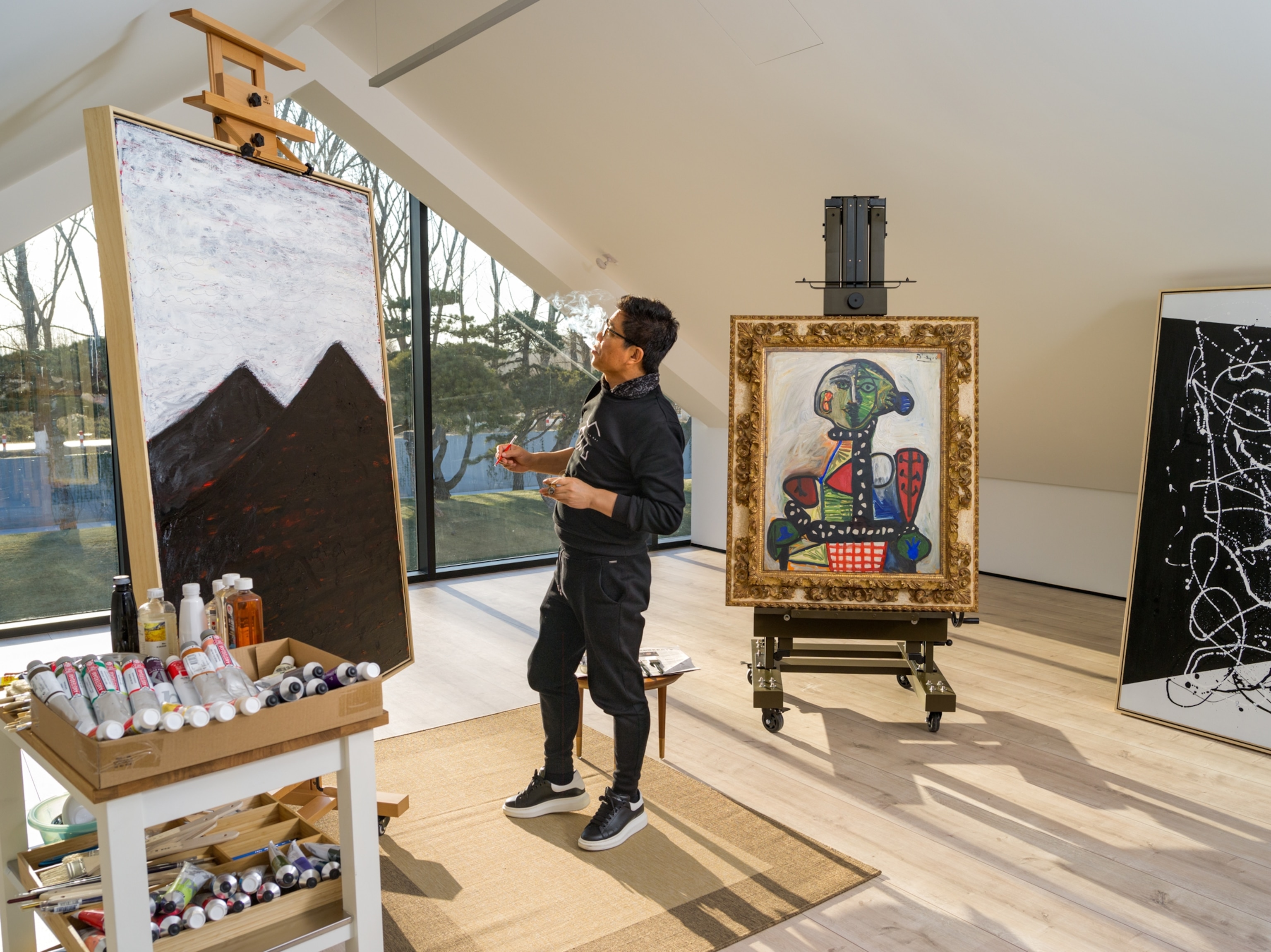 a Chinese film mogul standing and painting next to a Picasso painting on an easel