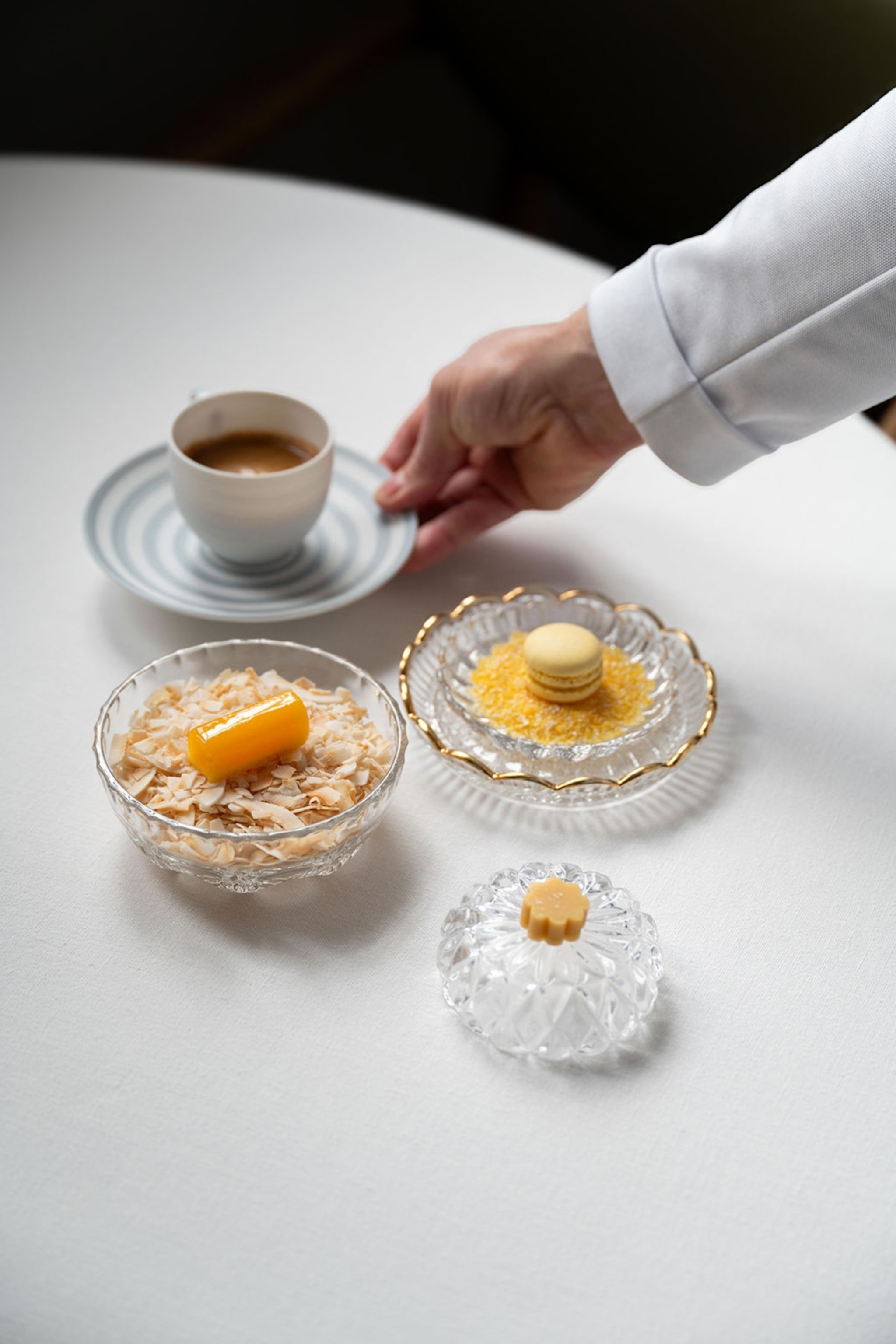 A sleek, linen-decked table with a hand placing down a cup of espresso next to small dessert places, including macarons.