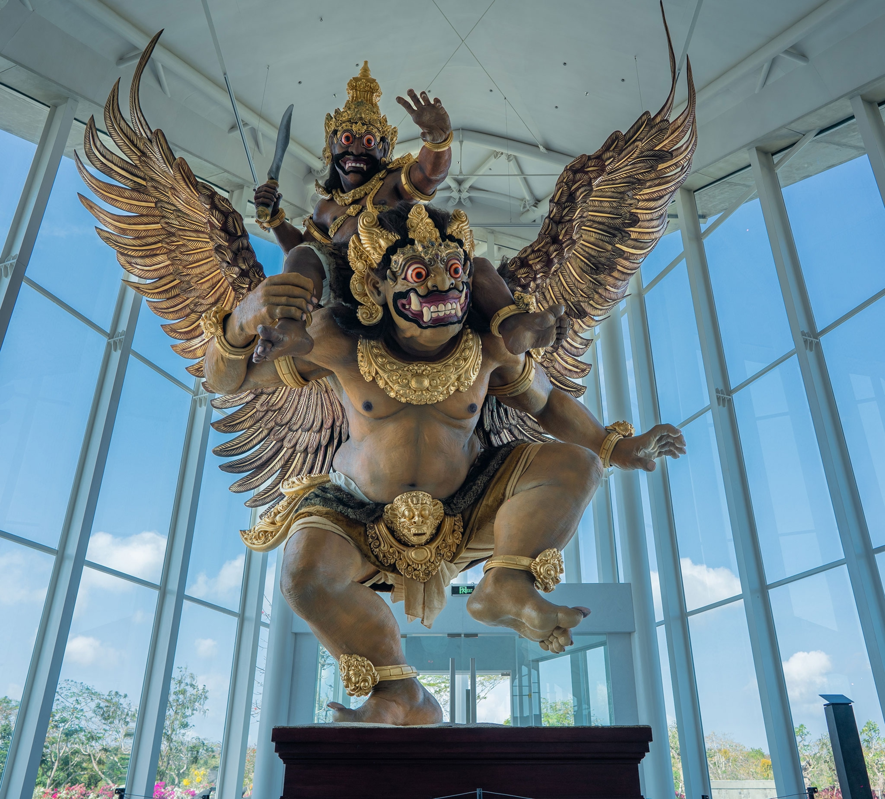 A statue of a man, naked except for ornate, gold anklets, an oranate and gold sash around his waste, gold bracelets and necklace. The man is wearing an ornate mask and has large, gold wings. Another man, dressed in the same way, with a knife, rides on his back.