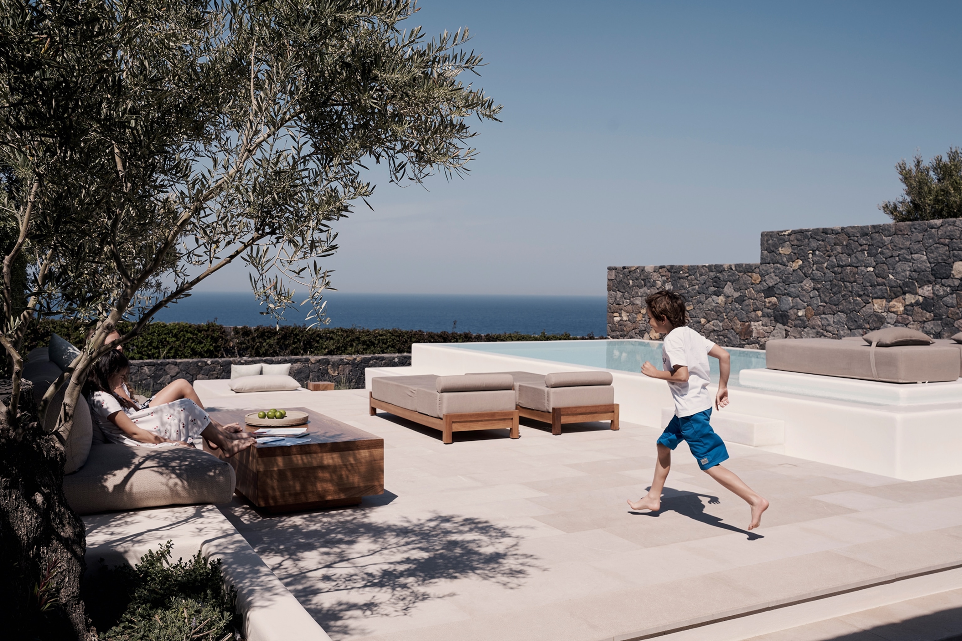 A pool facing the ocean as a boy runs to his mother, who is lounging under palm trees to the side.