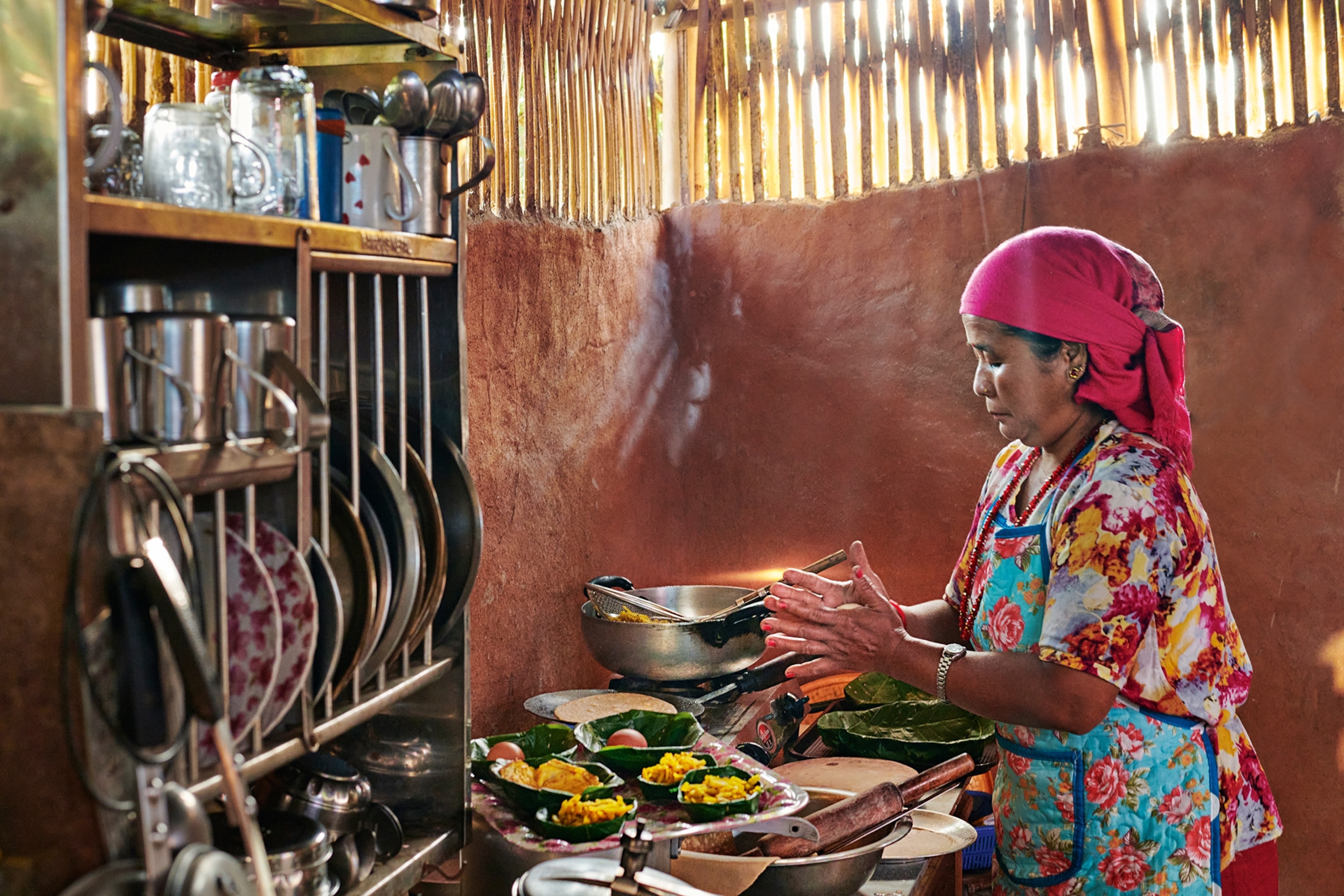 A wide shot of a simple mud-walled kitchen with a shelving system for plates, utensils and tins, as a woman prepares multiple dishes of food.