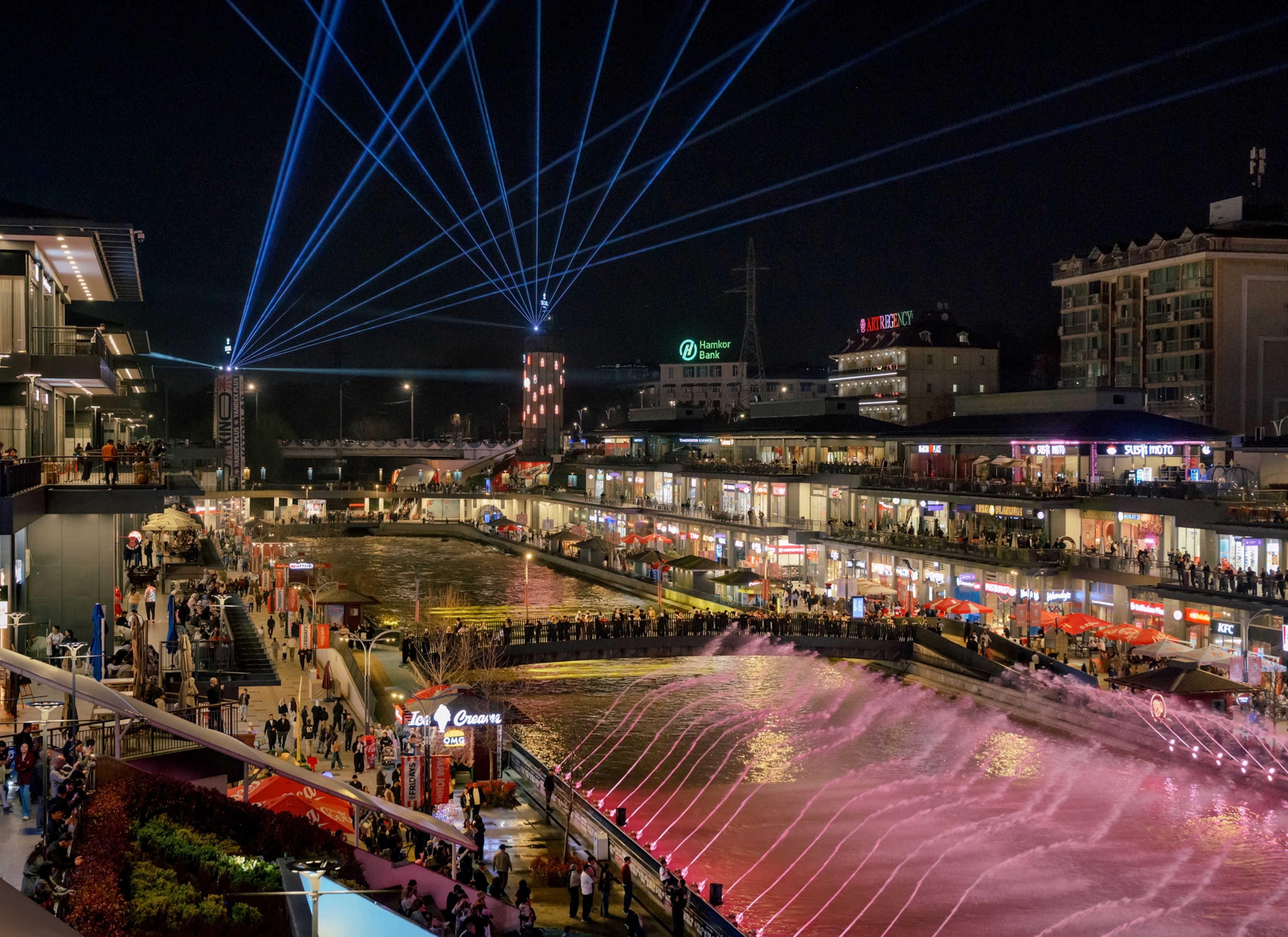 A cityscape with a large, man-made body of water sitting in the middle of a rectangular shopping center with various stores and restaurants. It is nighttime and a laser show displays above the water.