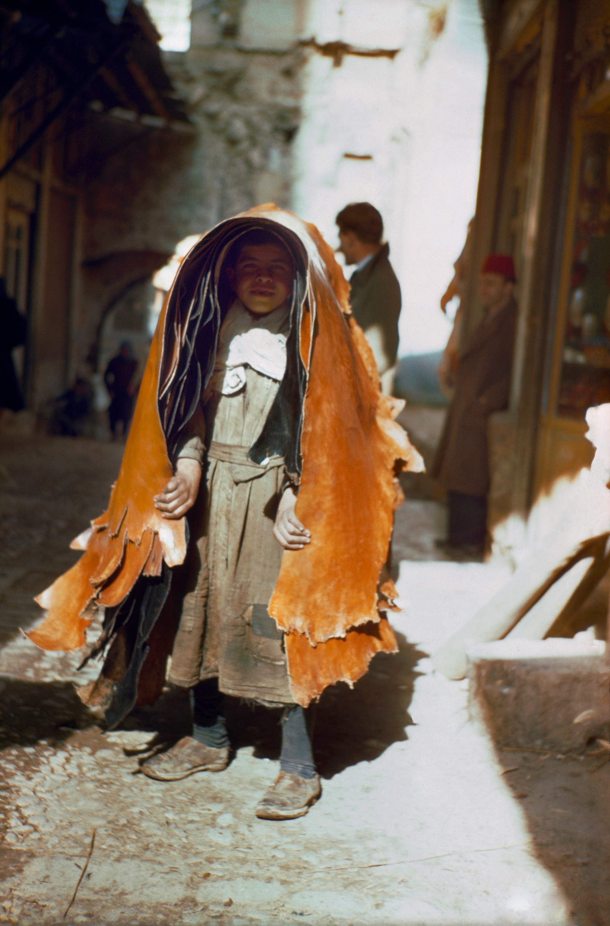 A young boy carries several pieces of hide through a narrow cobbled street, 1946