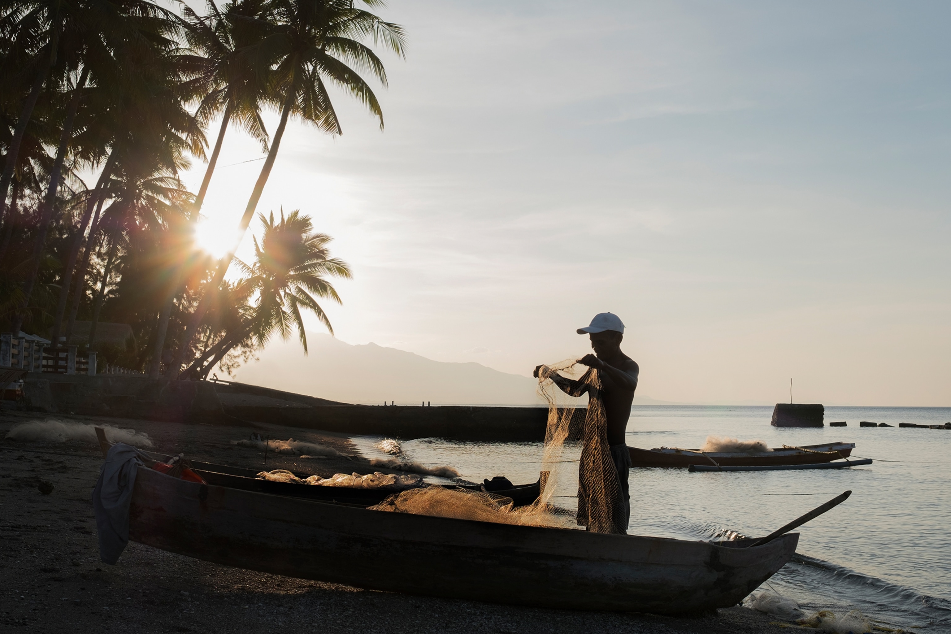 A local man unwinding a fishing net next to a boat on a tropical beach at sunset.