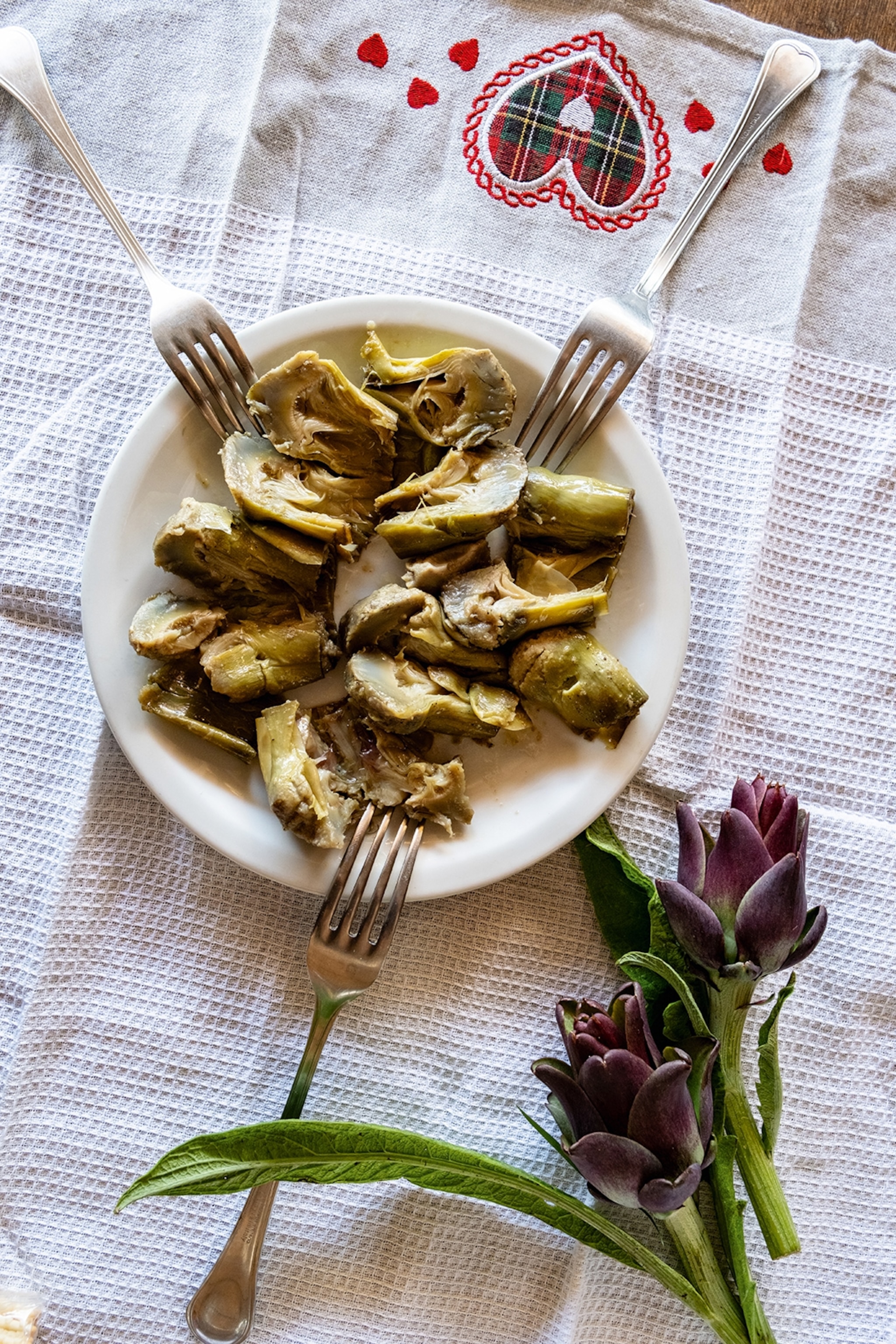 A plate of cooked artichokes with three forks on a rural tablecloth with fresh artichokes on the side.