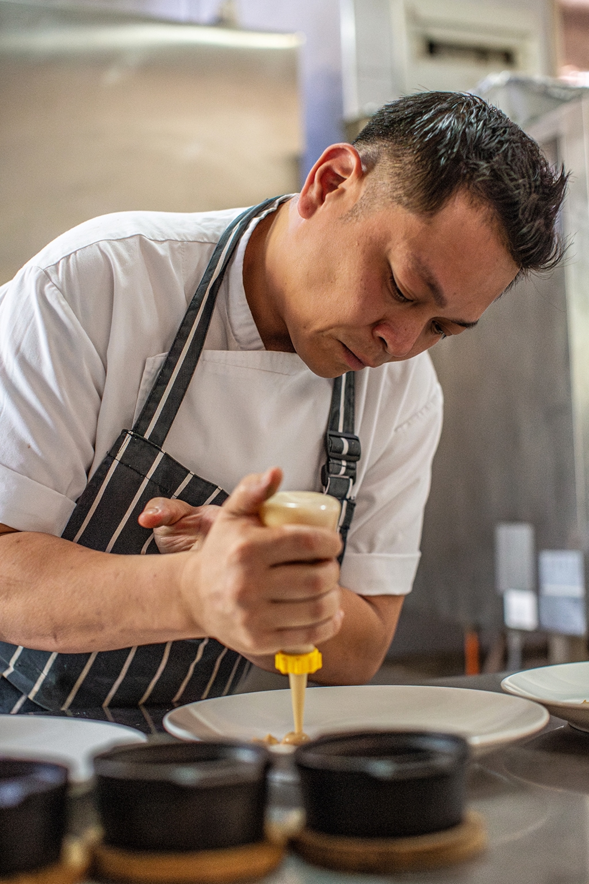 A male chef wearing an apron in a kitchen, leaning over a counter to squeeze sauce onto a plate.