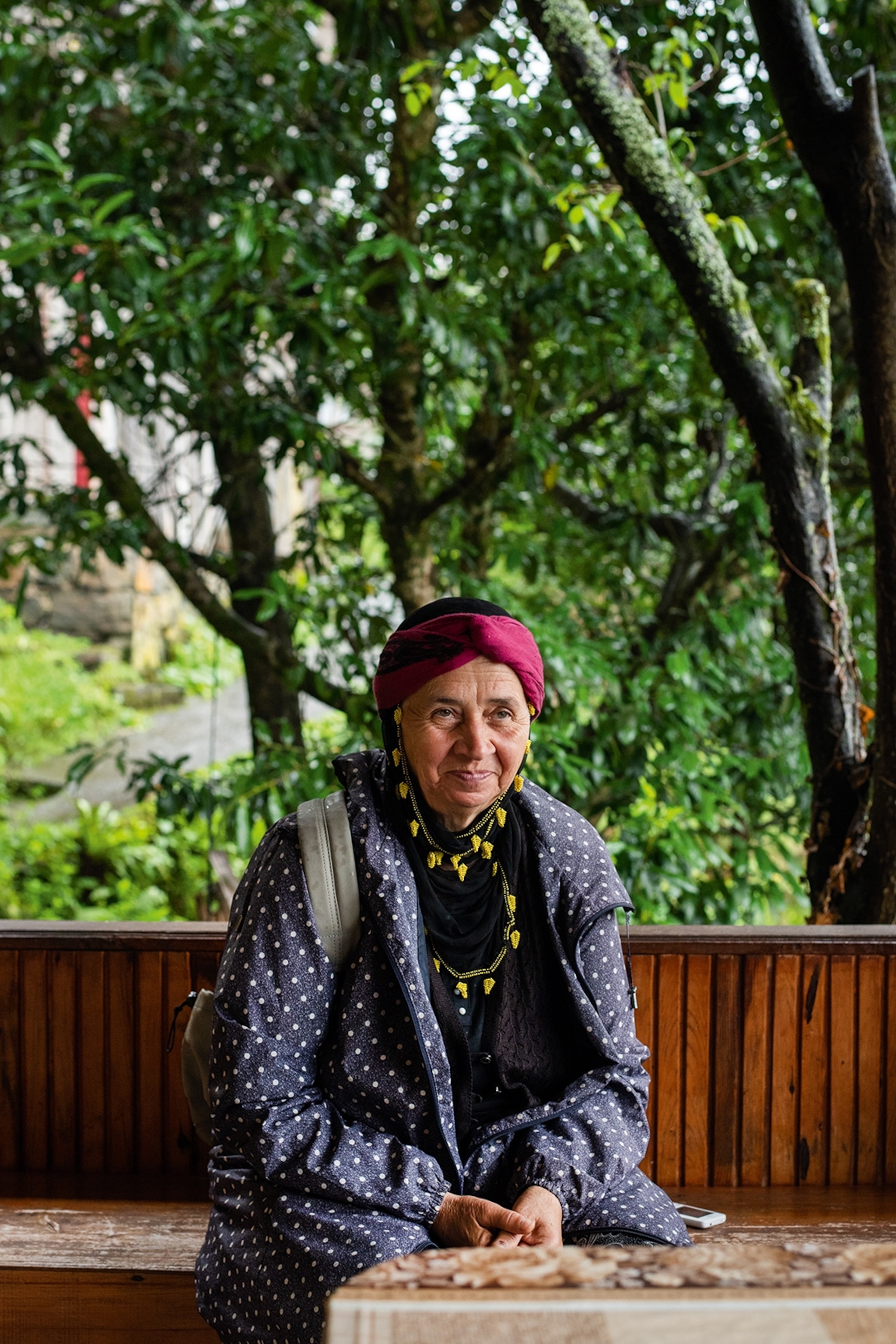 An elderly woman smiling and sitting on a wooden bench with lush trees in the background.