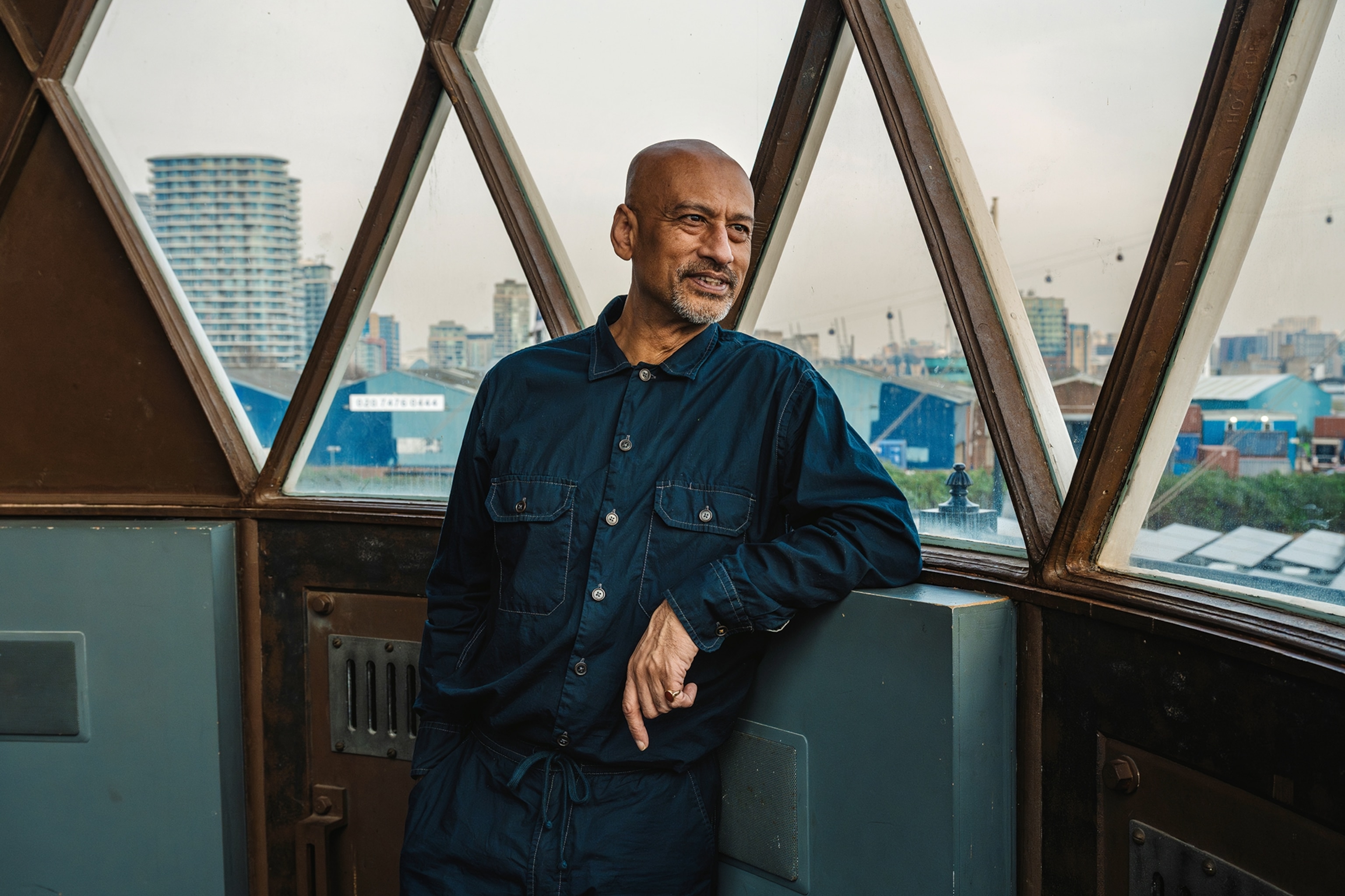 A middle-aged man in a blue overall looking out the window of an urban lighthouse.