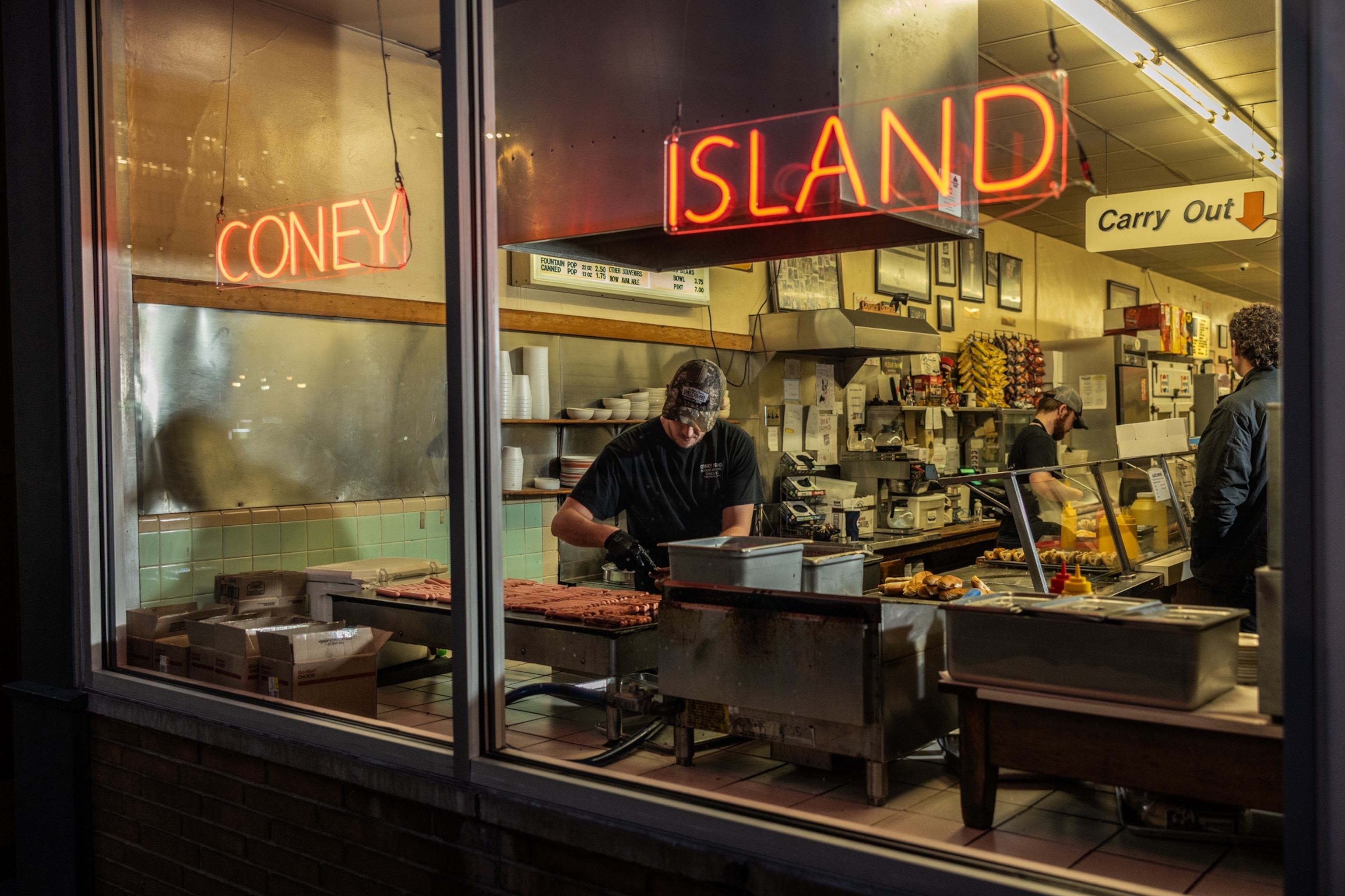 A view through the window as two workers make hot dogs and a man stands at the counter observing.