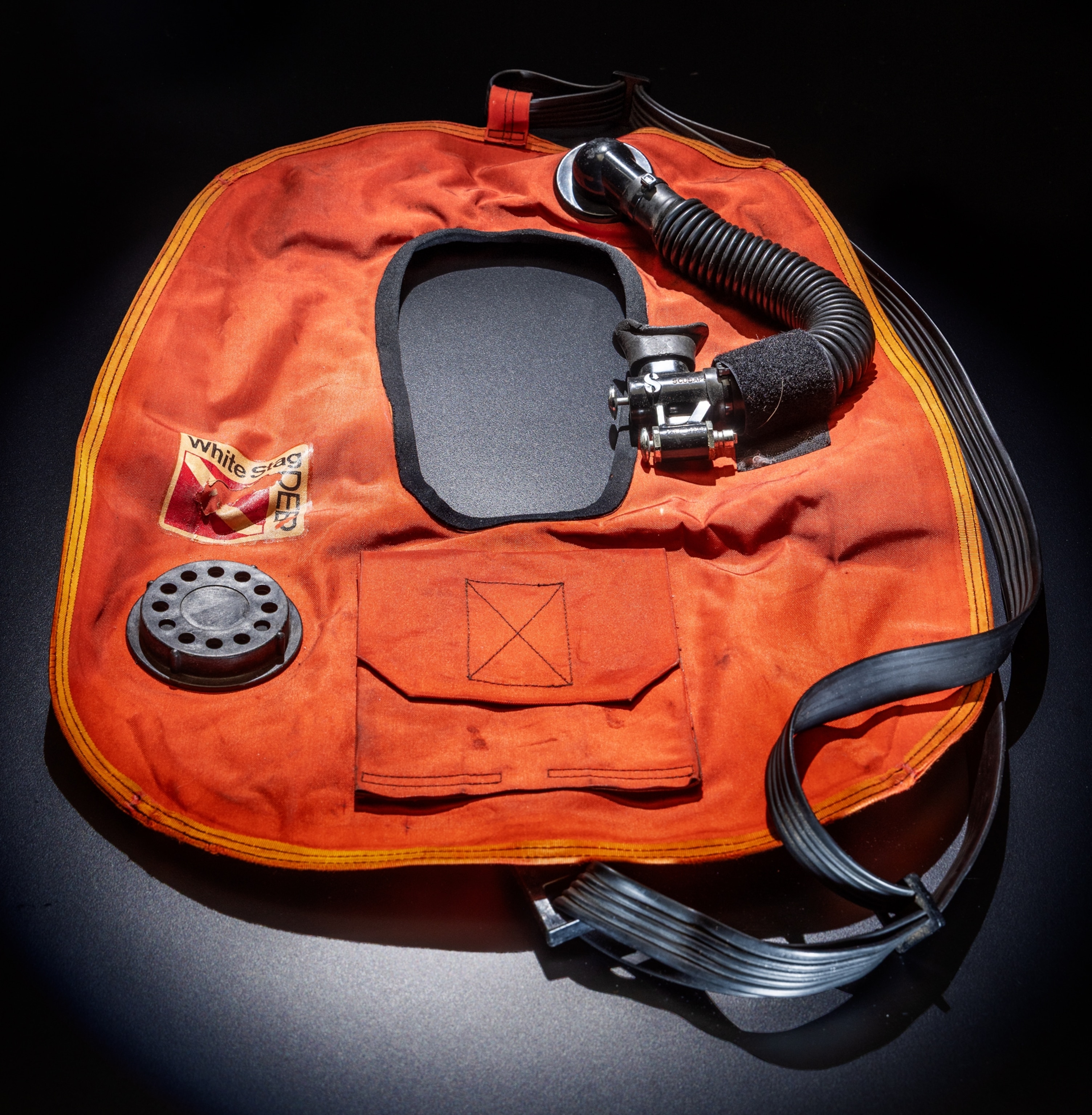 An oval orange vest with a head hole cut out in the middle. On the front of it is a pocket, air tube, and one long strap connected to both ends.