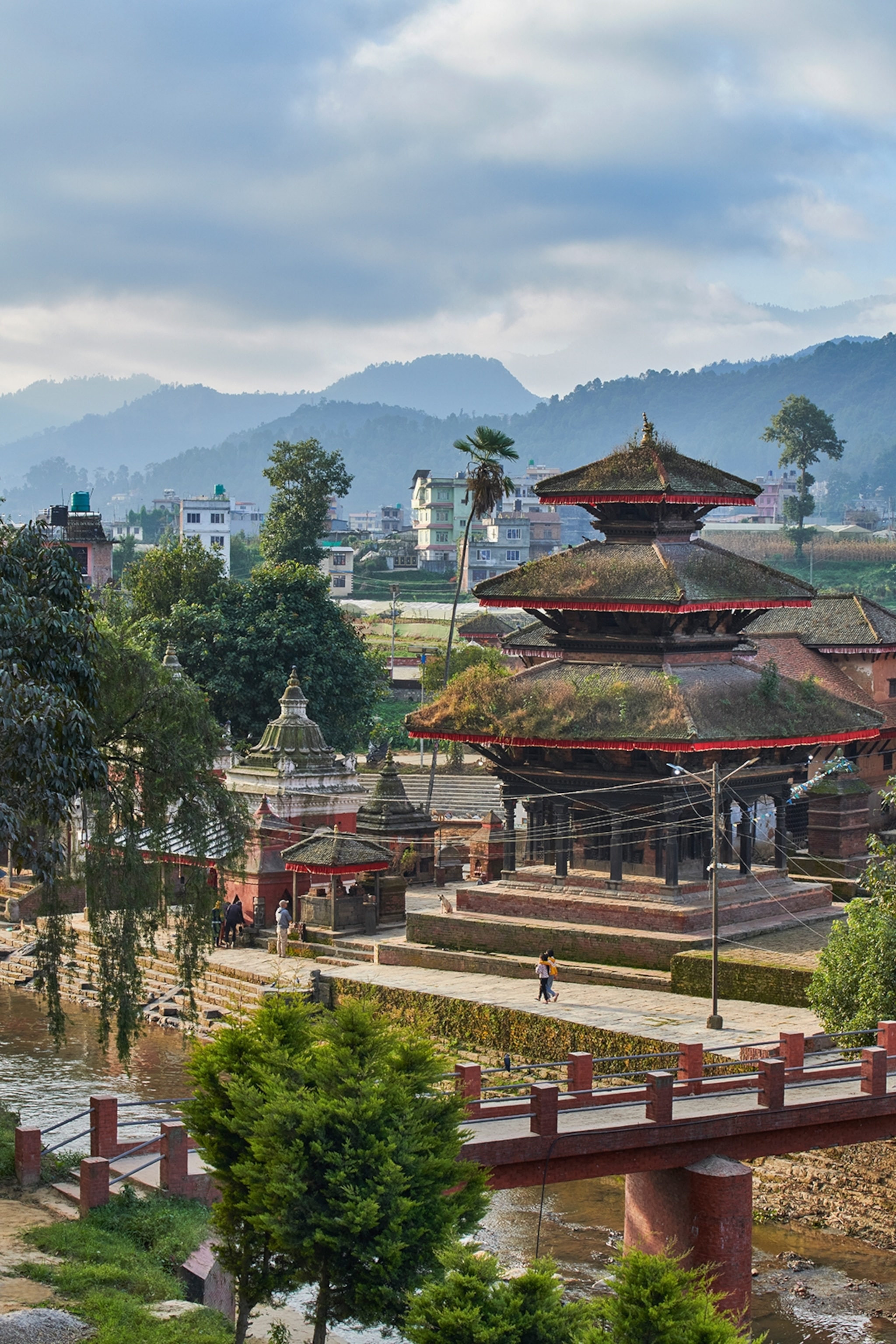 A low-level, ancient town with a moss-roofed temple, a river running by and mountains in the background.