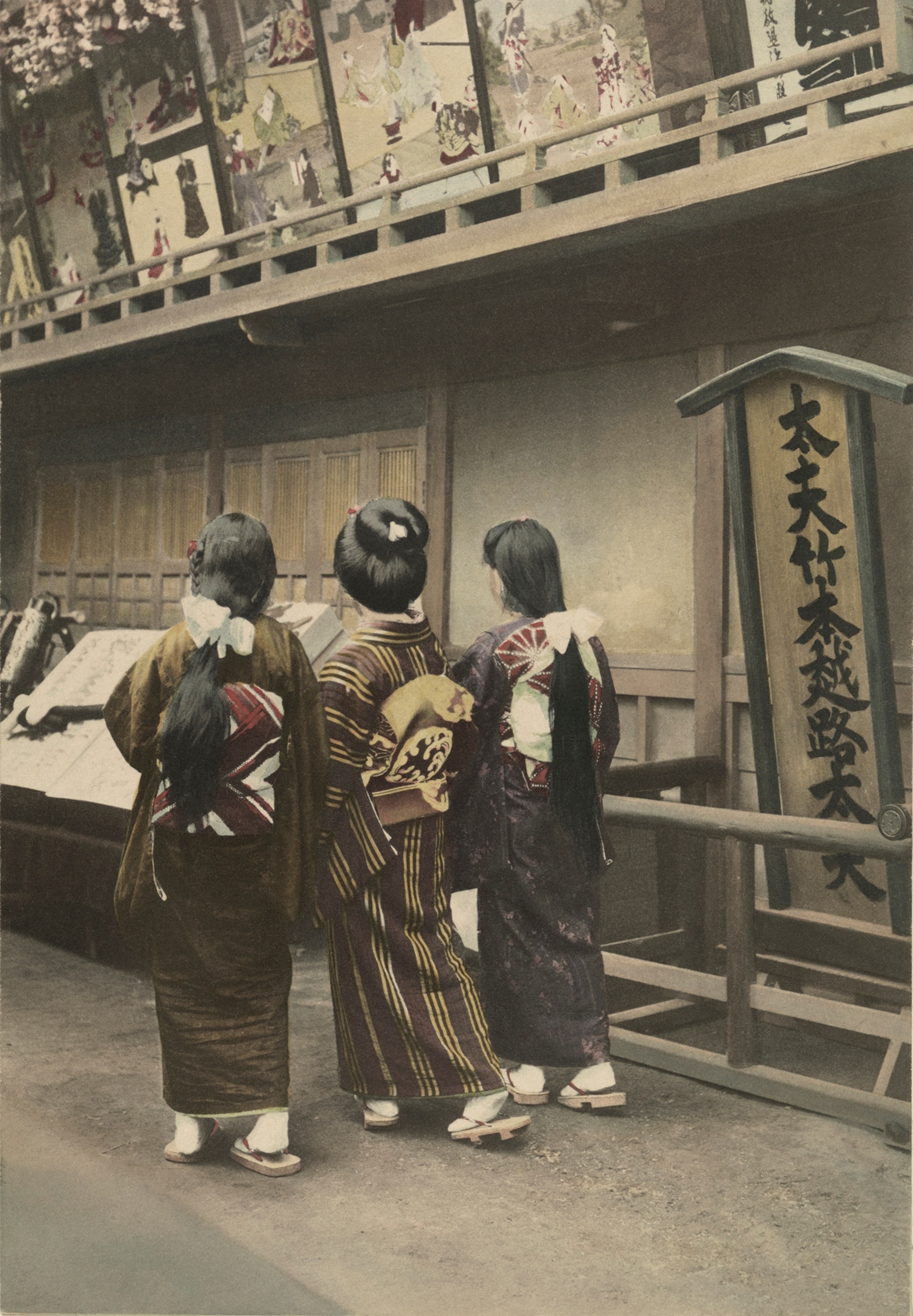 Three women walk to a matinee show at the theater. They are on their way to a performance advertised by the hand-drawn posters seen at the top of the illustration, and the name of the star, Tayu Takemoto Koshiji, is blazoned not he panel at the right.